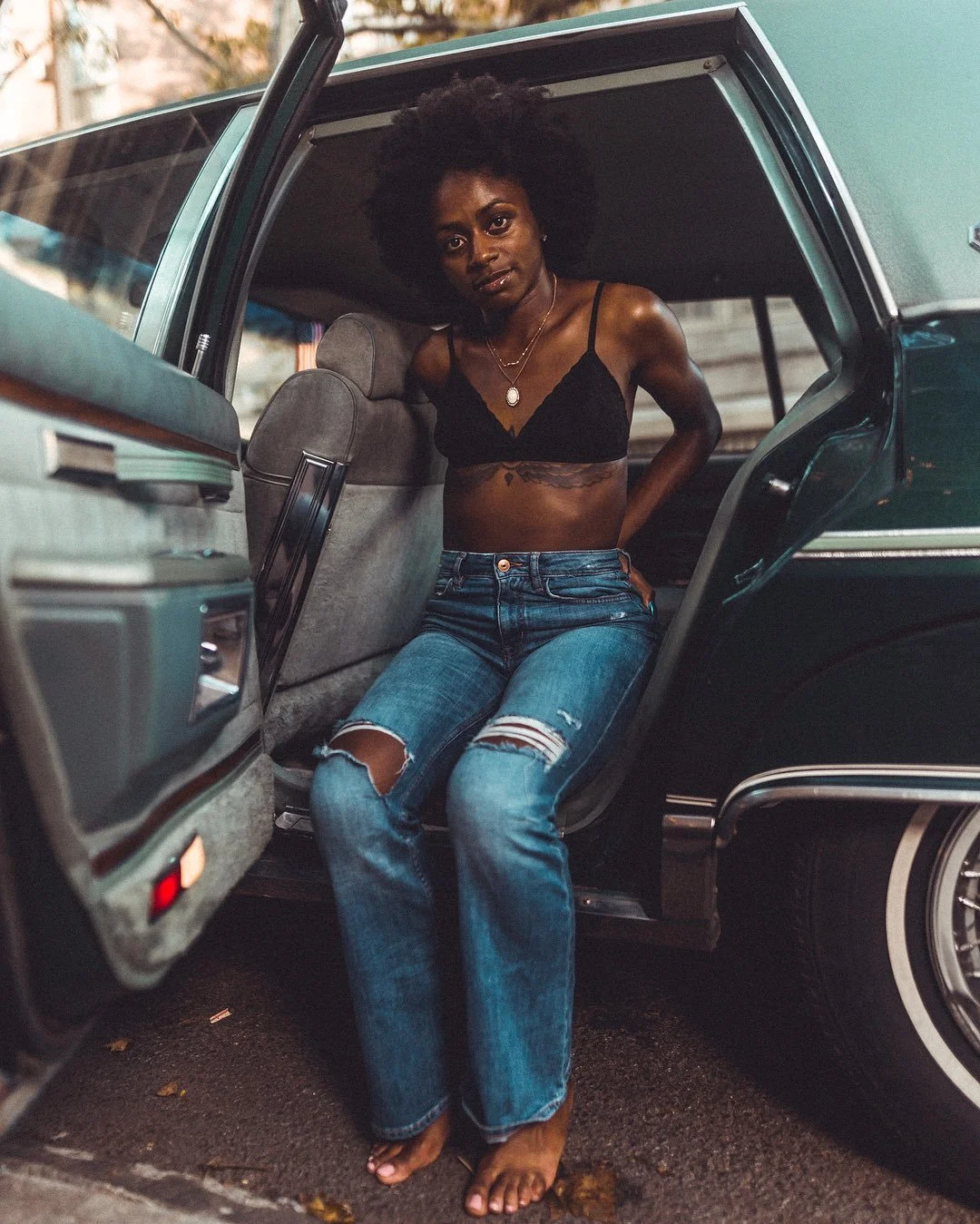 A woman with curly hair, wearing a black bra, ripped jeans, and sandals, is standing at the open door of a vintage green car, looking at the camera with a serious expression.