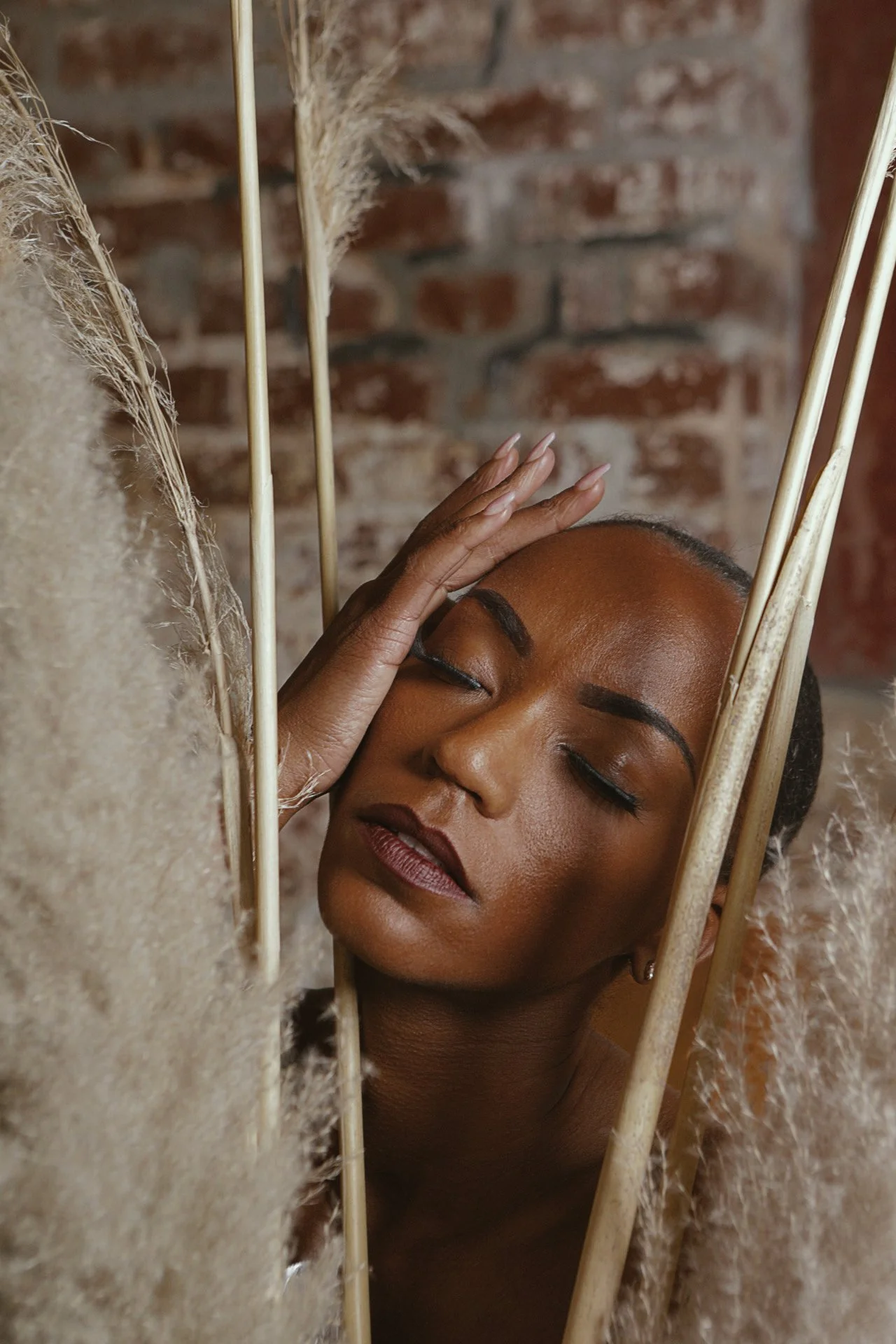 A woman with closed eyes and crossed arms resting her head against a mirror, surrounded by tall dried pampas grass and wooden sticks, with a brick wall in the background.