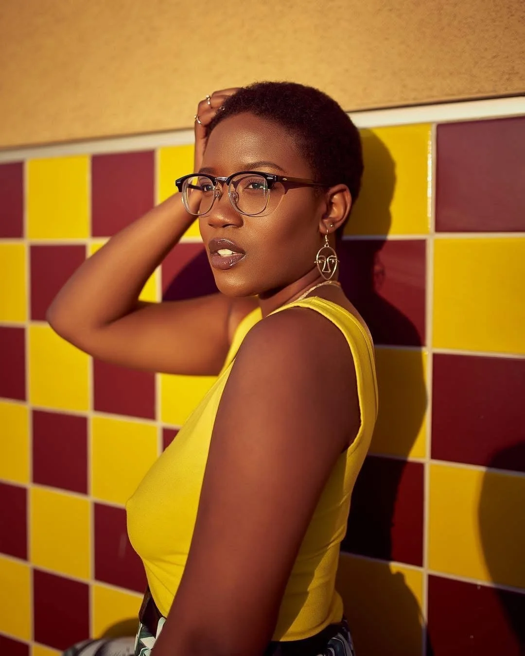 A woman with short hair wearing glasses, earrings, and a yellow sleeveless top, posing against a yellow and red checkered wall.
