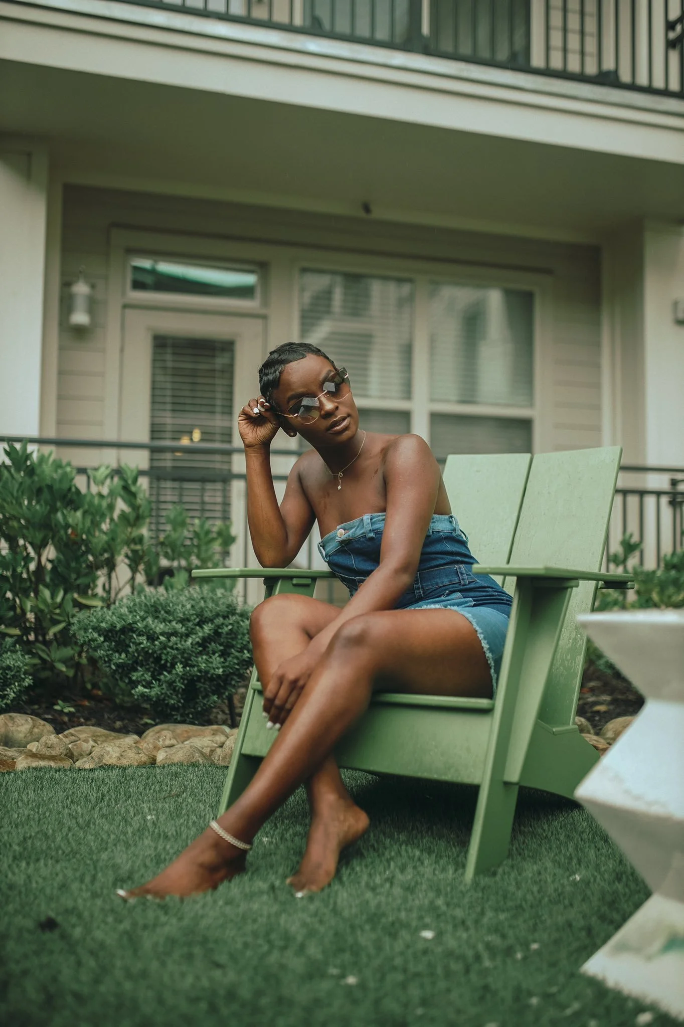 A woman with dark skin and short hair wearing sunglasses, a strapless denim dress, and ankle jewelry sitting outdoors on a green chair in a garden area with a house in the background.