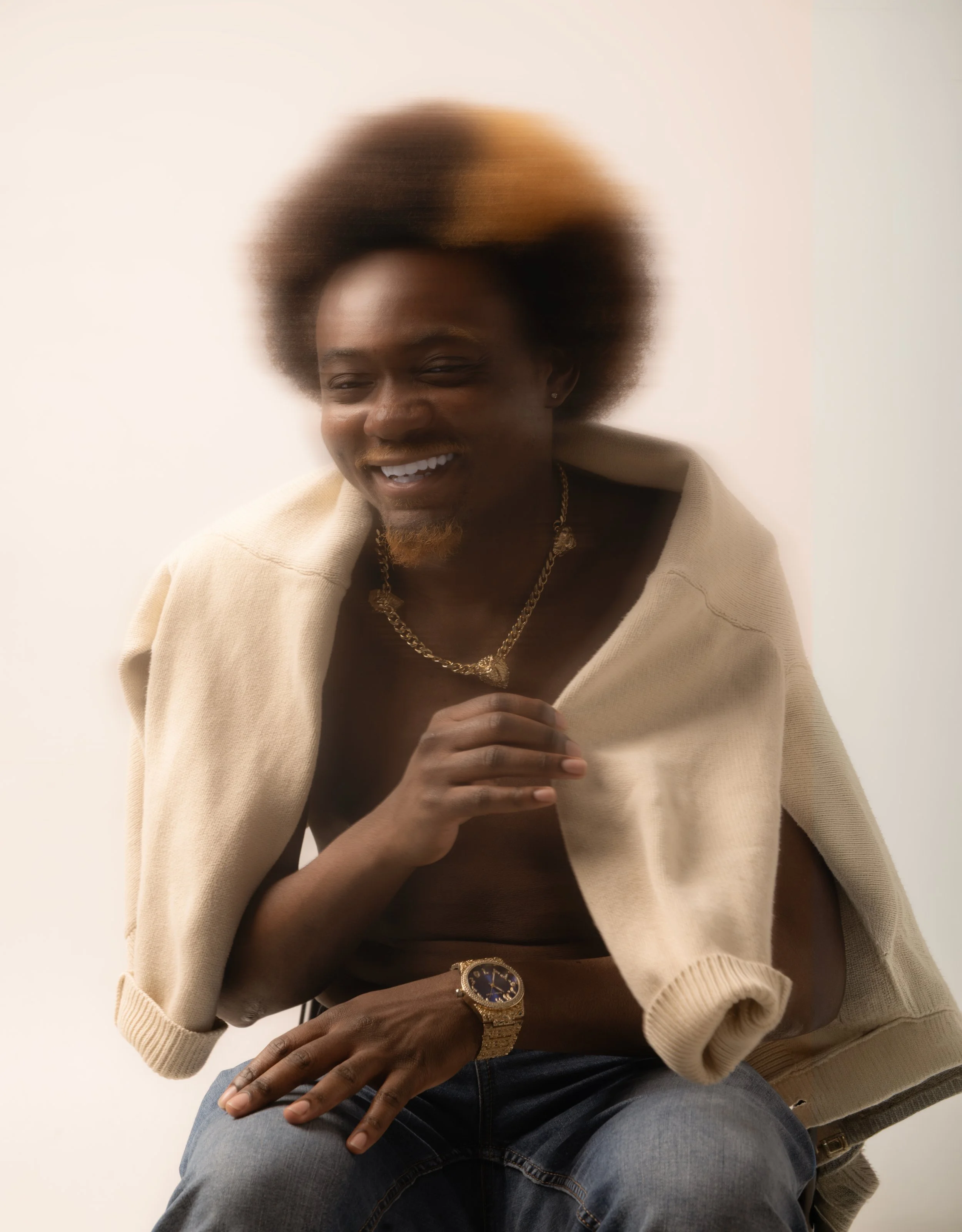 A smiling man with an afro hairstyle, wearing a gold watch, gold necklace, jeans, and a cream-colored sweater draped over his shoulders against a plain background.