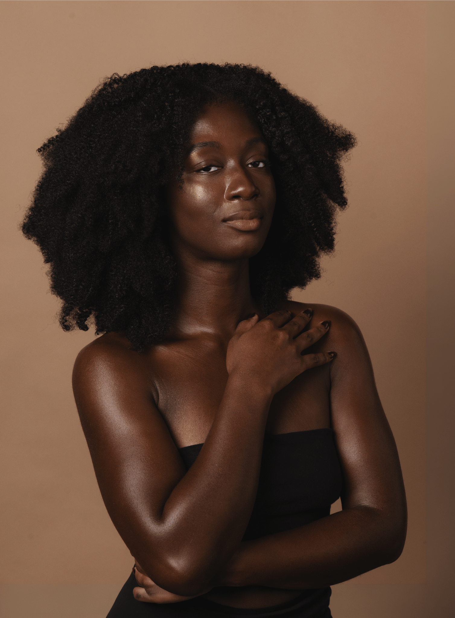 A portrait of a young Black woman with natural curly hair, wearing a black strapless top, and posing with her hand on her shoulder against a neutral background.