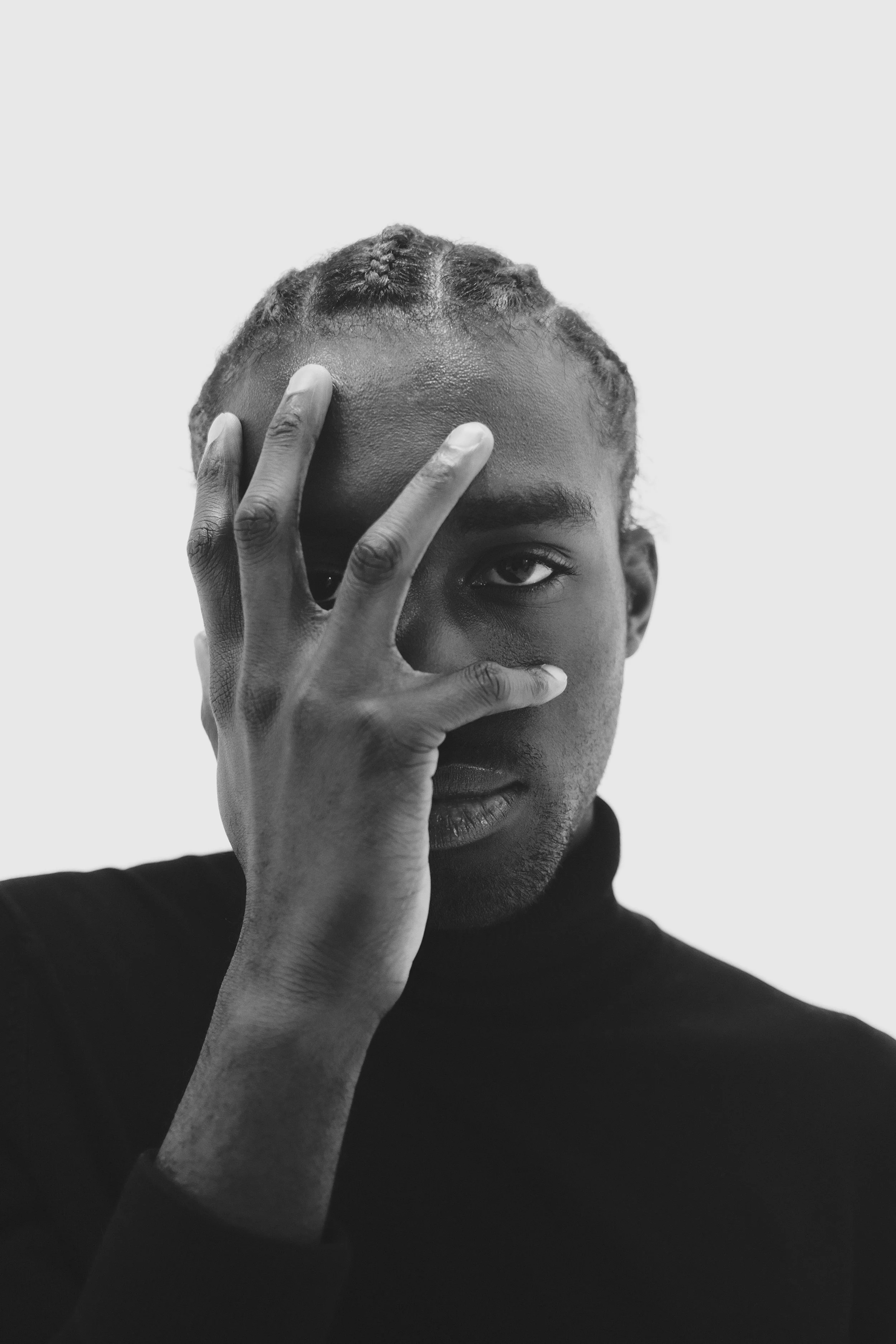 A black-and-white portrait of a person with braided hair, covering one eye with their hand, and looking directly at the camera.