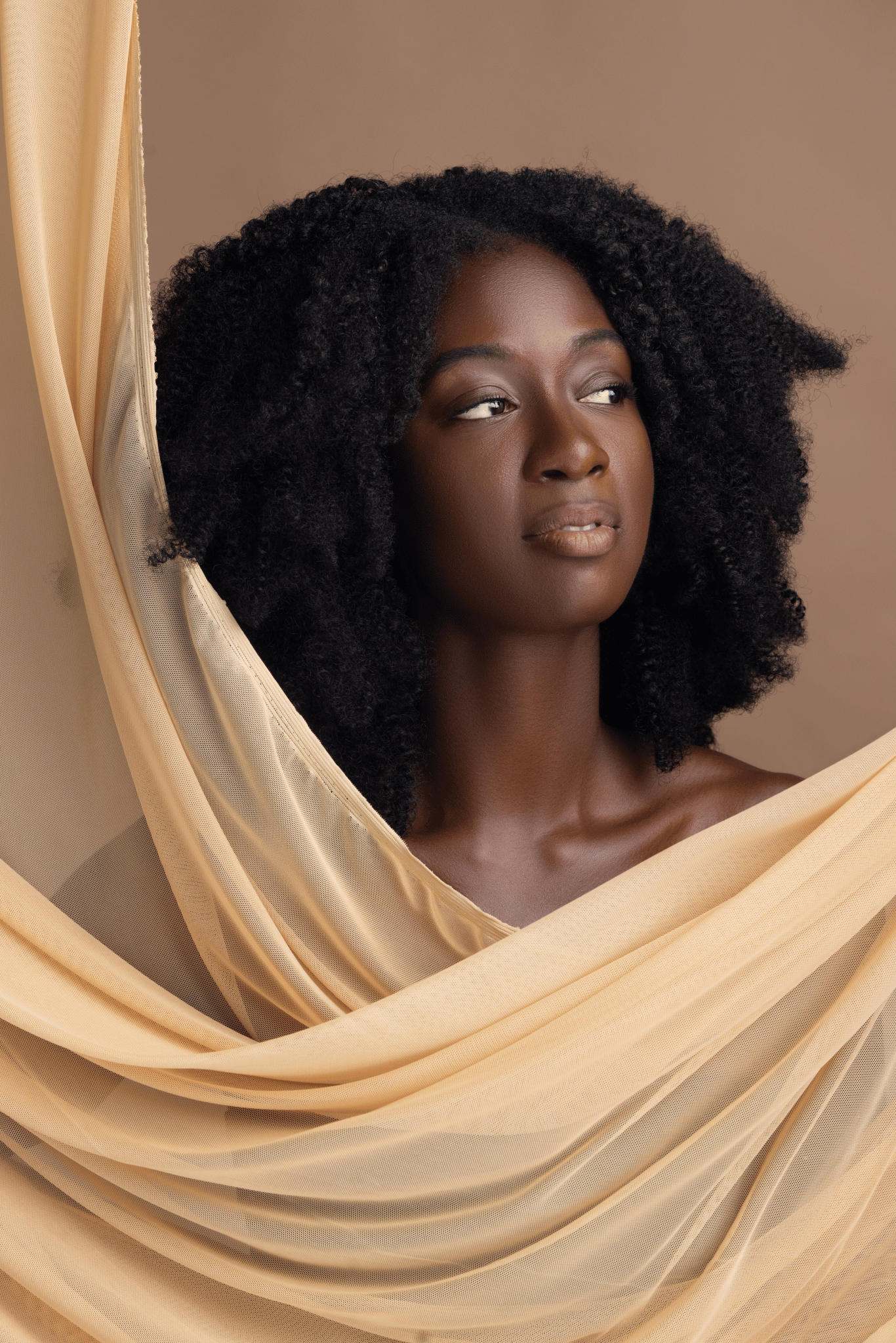 Portrait of a woman with natural curly hair, gazing to the side, framed by beige draped fabric.