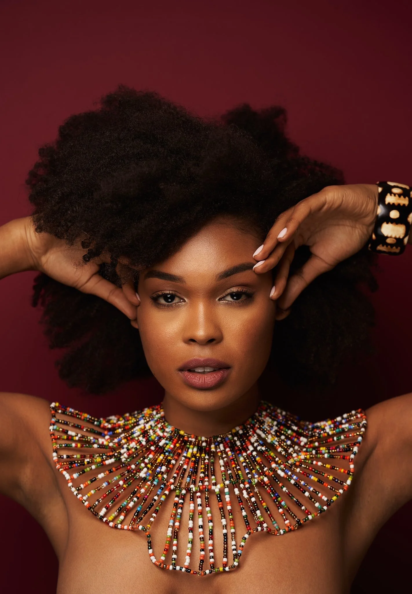 A woman with natural hair lying down against a dark red background, wearing a colorful beaded necklace and a black bracelet.