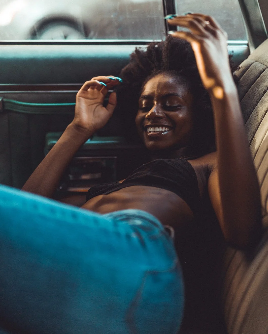 A young woman with curly hair and dark skin lying across the backseat of a car, smiling with eyes closed, wearing a black top and blue jeans, with one arm raised and the other touching her hair.