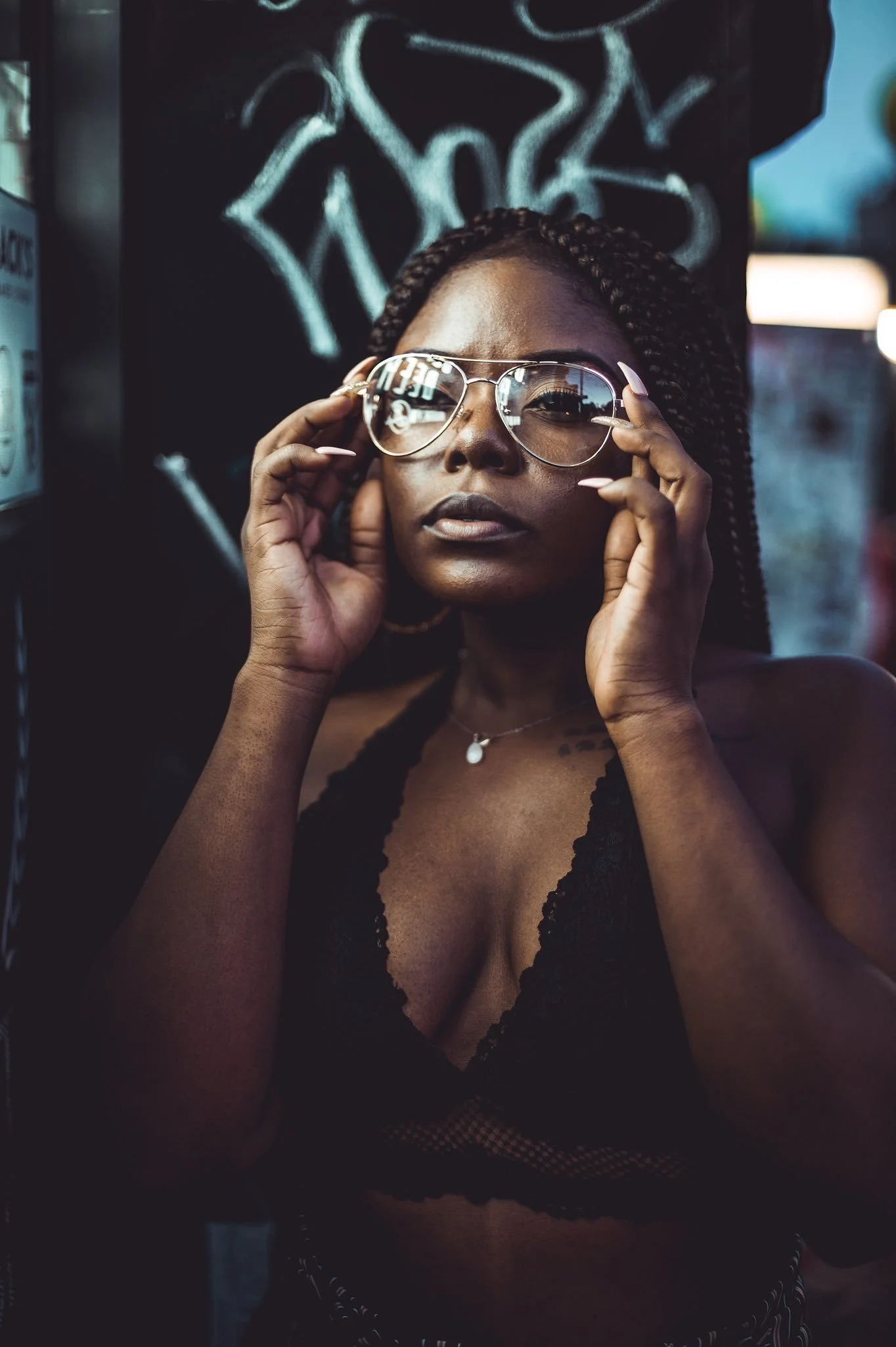 A woman with dark skin and braided hair wearing glasses, a black lace top, and a necklace, adjusting her sunglasses while looking directly at the camera.