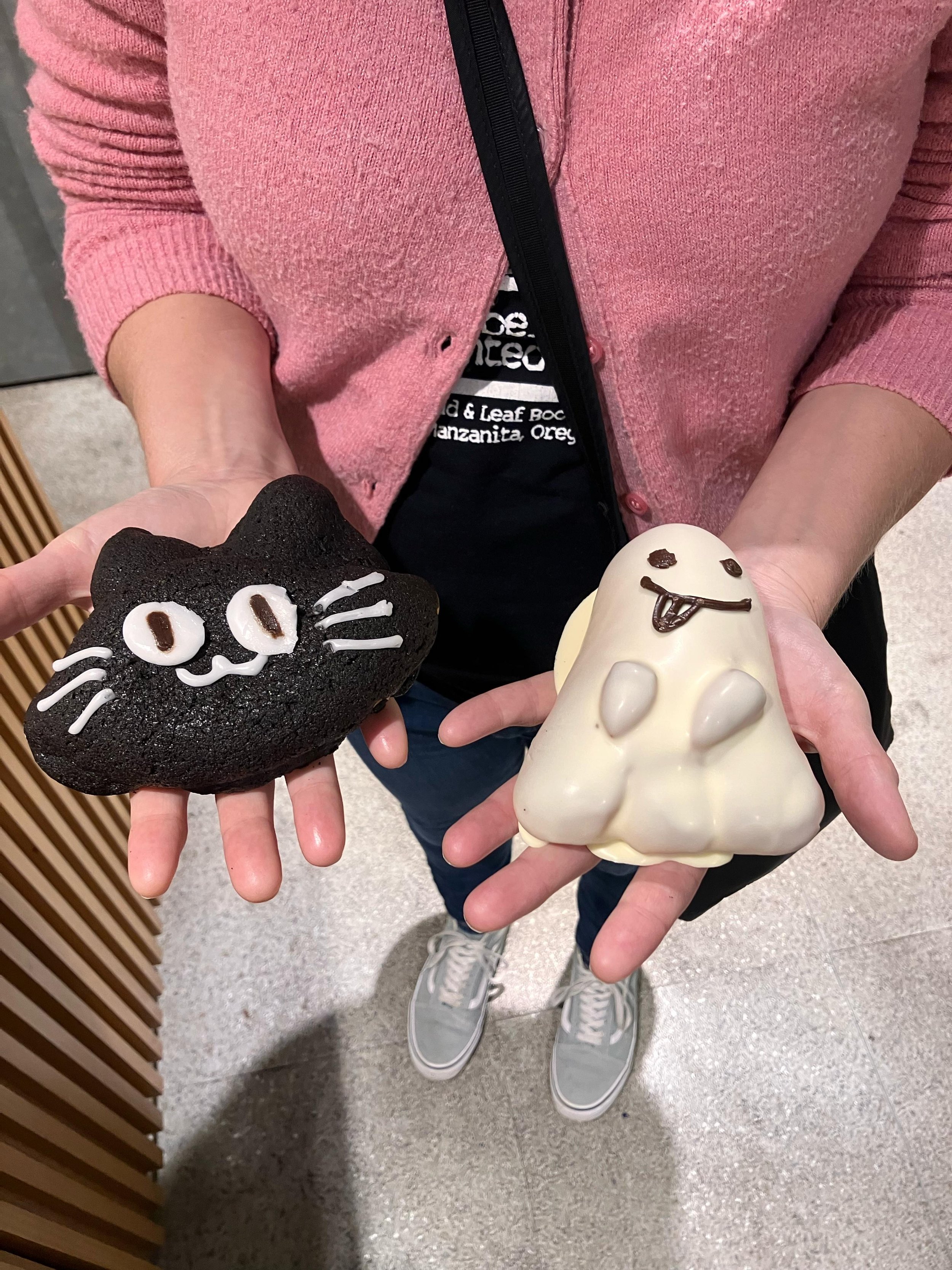 Person holding two Halloween-themed cookies, one black cat and one ghost, decorated with frosting and face details.