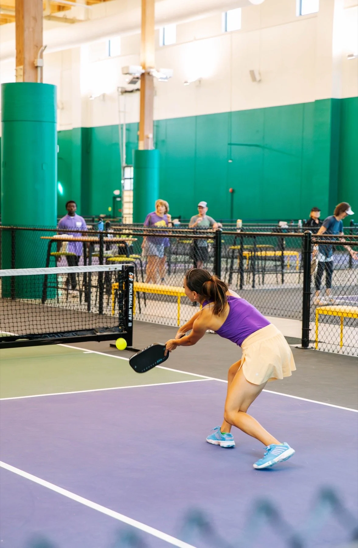 A woman playing pickleball on an indoor court, preparing to hit a yellow ball with a black paddle, with other players and spectators in the background.