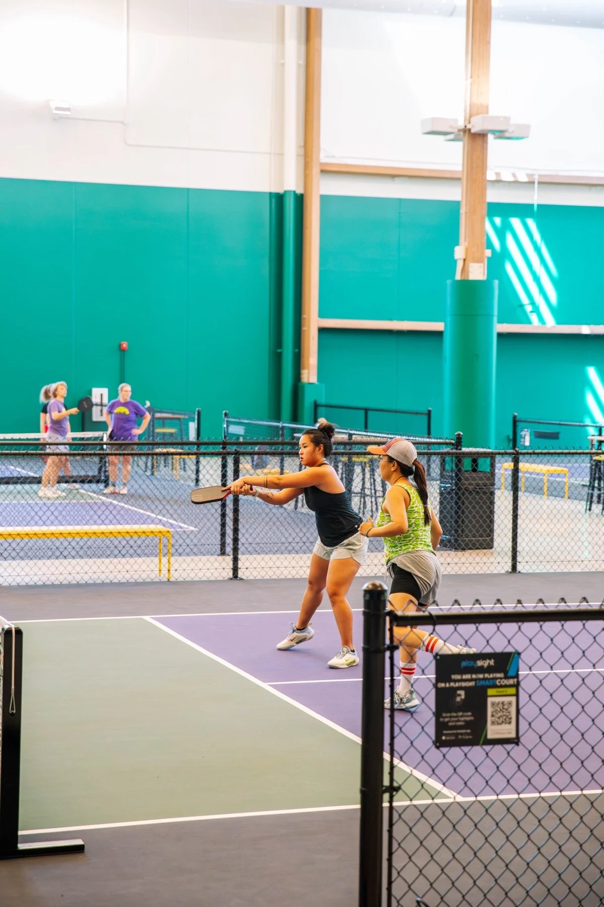 Two women playing pickleball indoors, with others in the background near a ping pong table.