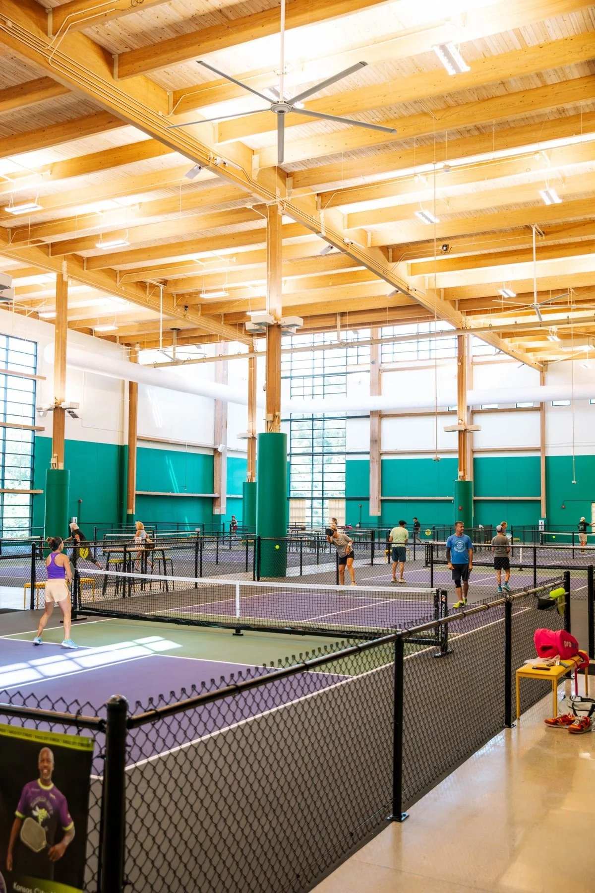 Indoor pickleball courts with multiple playing areas, players playing, and spectators watching, in a large wooden-beamed gymnasium.