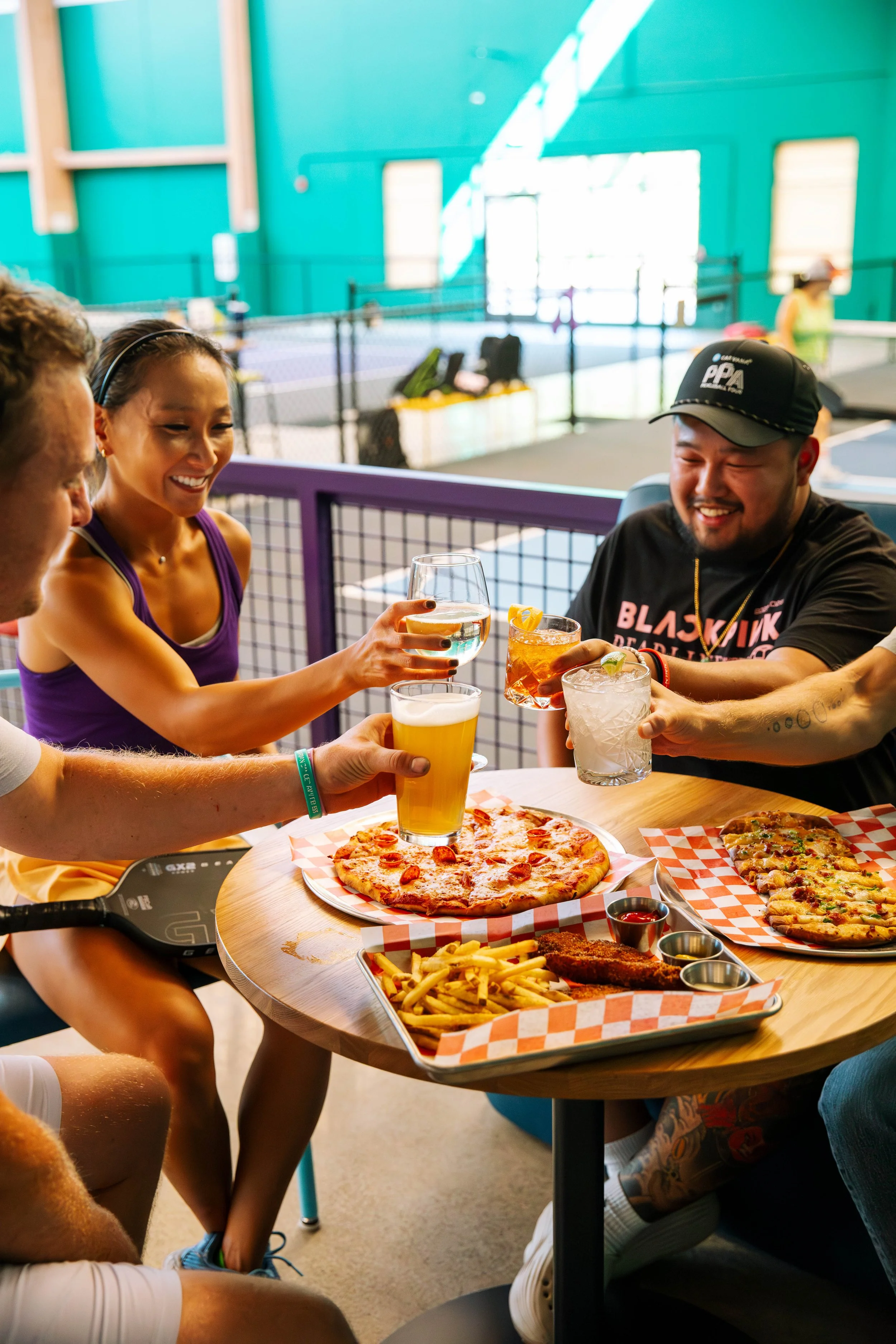 Group of four friends enjoying drinks and pizza at a table in an indoor sports facility.