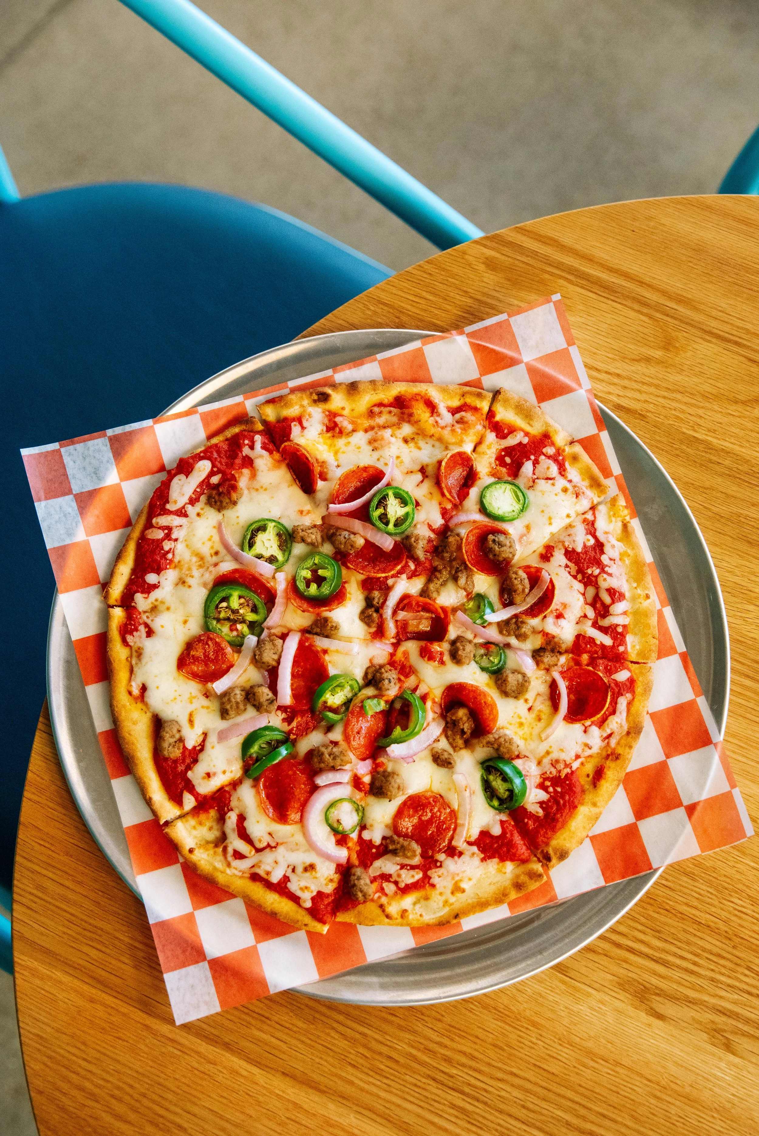 A cheese pizza topped with pepperoni, sausage, green jalapeno slices, and red onion on a checkered paper lining, placed on a metal tray on a wooden table.