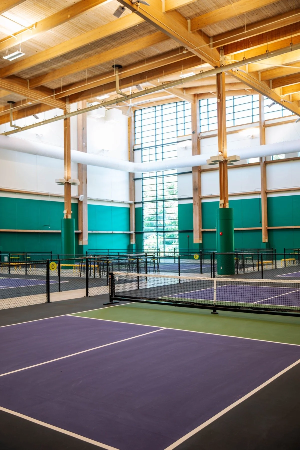 Indoor pickleball facility with purple and green courts, black fences, wooden beams, and large windows.