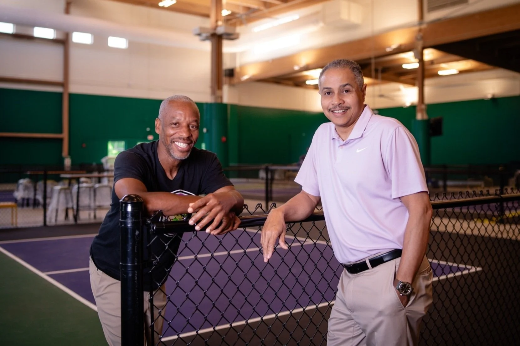 Two men smiling and leaning on a metal fence at an indoor pickleball court