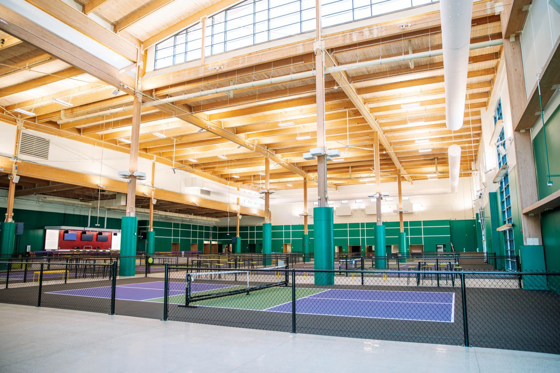 Indoor tennis courts with purple surfaces, surrounded by black fencing and green padding on the support columns, under a high wooden ceiling with large windows.