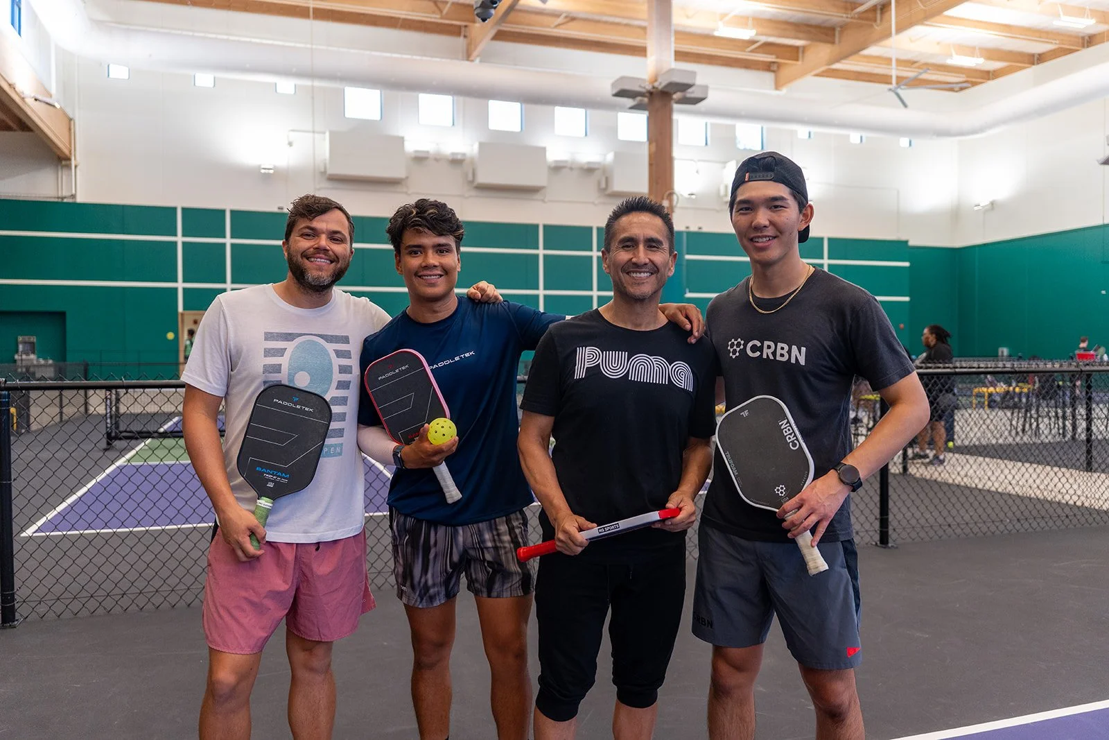 Four men standing on a pickleball court holding paddles and paddleball. They are smiling, with one man in the middle holding a paddledball. The court is indoors with a high wooden ceiling and a green wall with white lines.