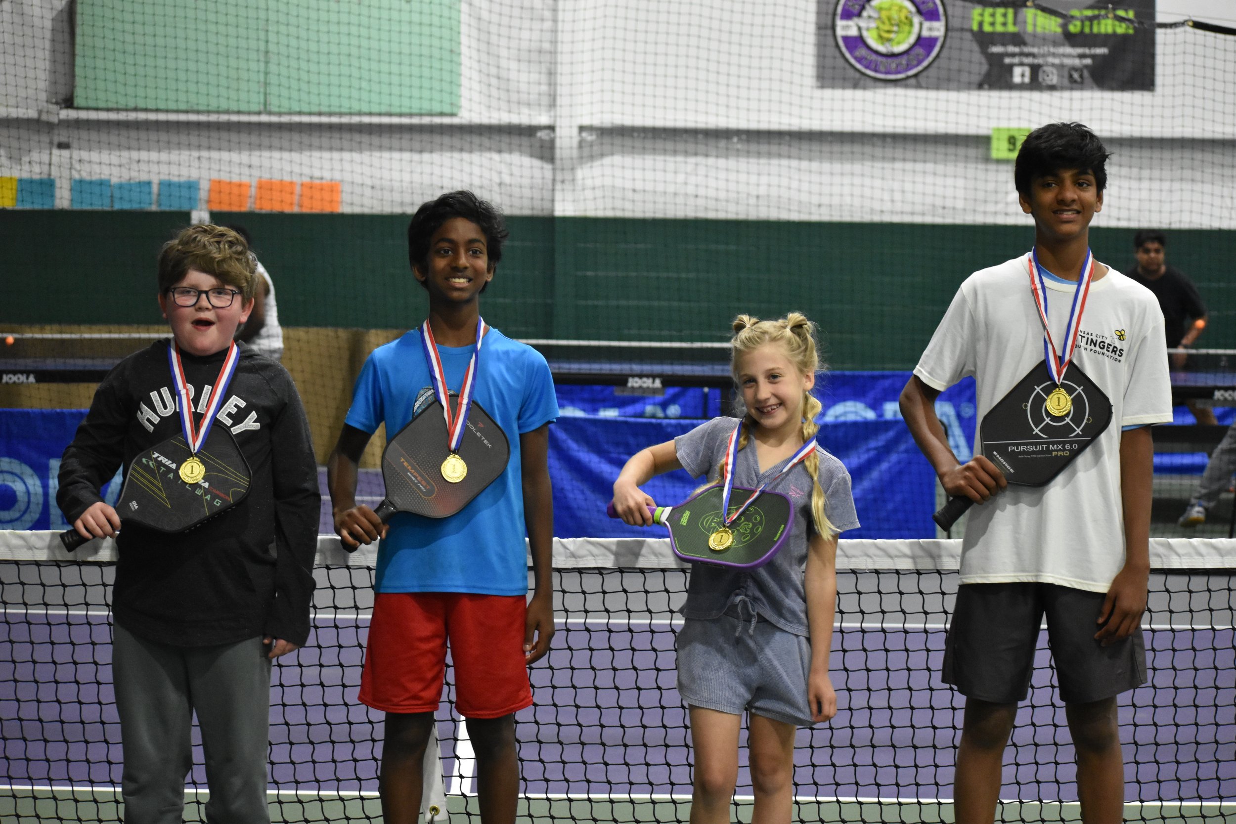Four children standing on a tennis court holding paddles and wearing medals after a match or tournament.