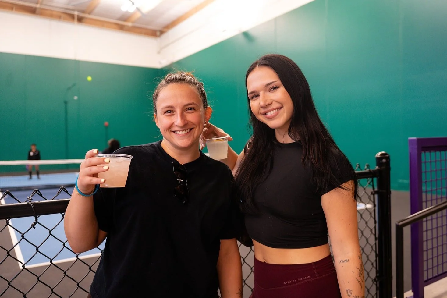 Two smiling women holding drinks at an indoor tennis court.