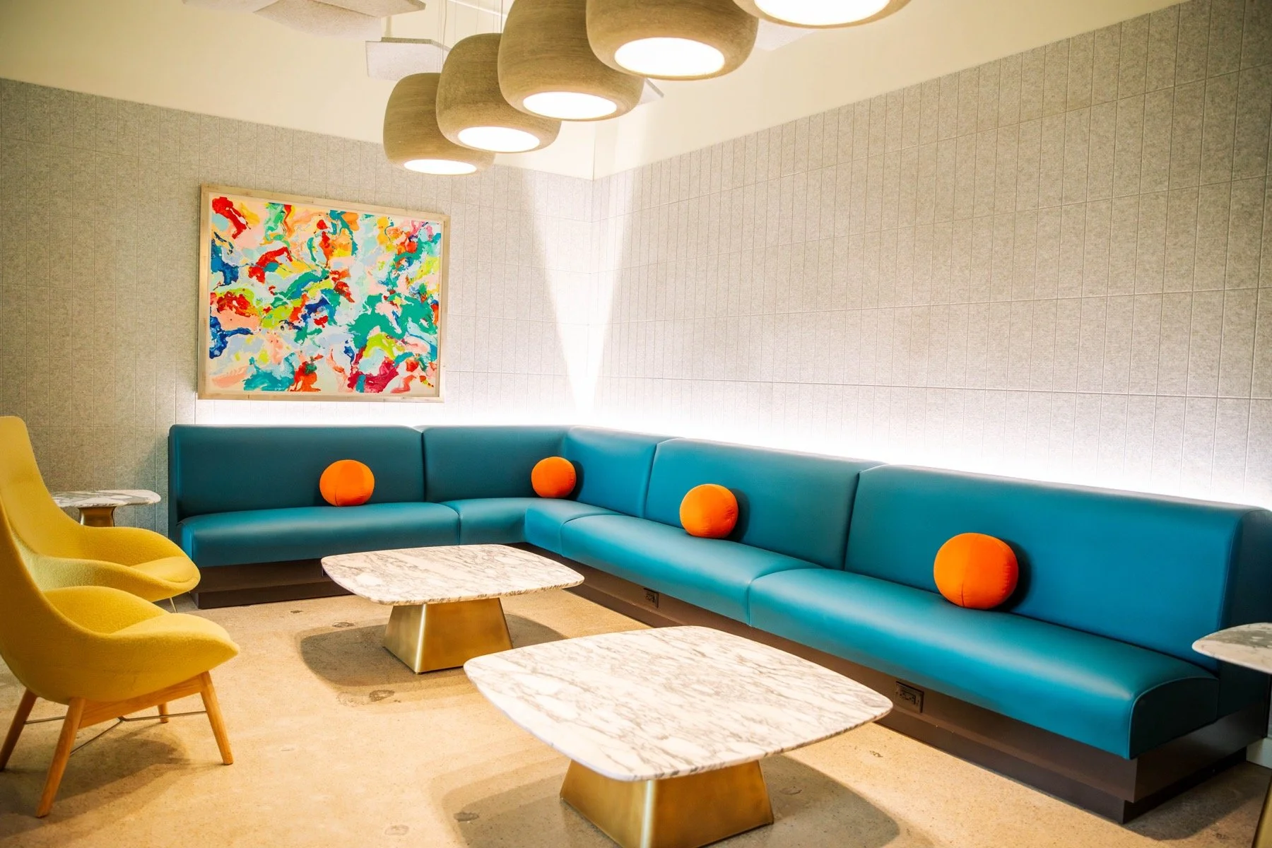 Modern lounge with a long blue sectional sofa decorated with four orange round pillows, yellow armchairs, three marble-top coffee tables, a colorful abstract painting, and a ceiling light fixture with multiple beige hanging shades.