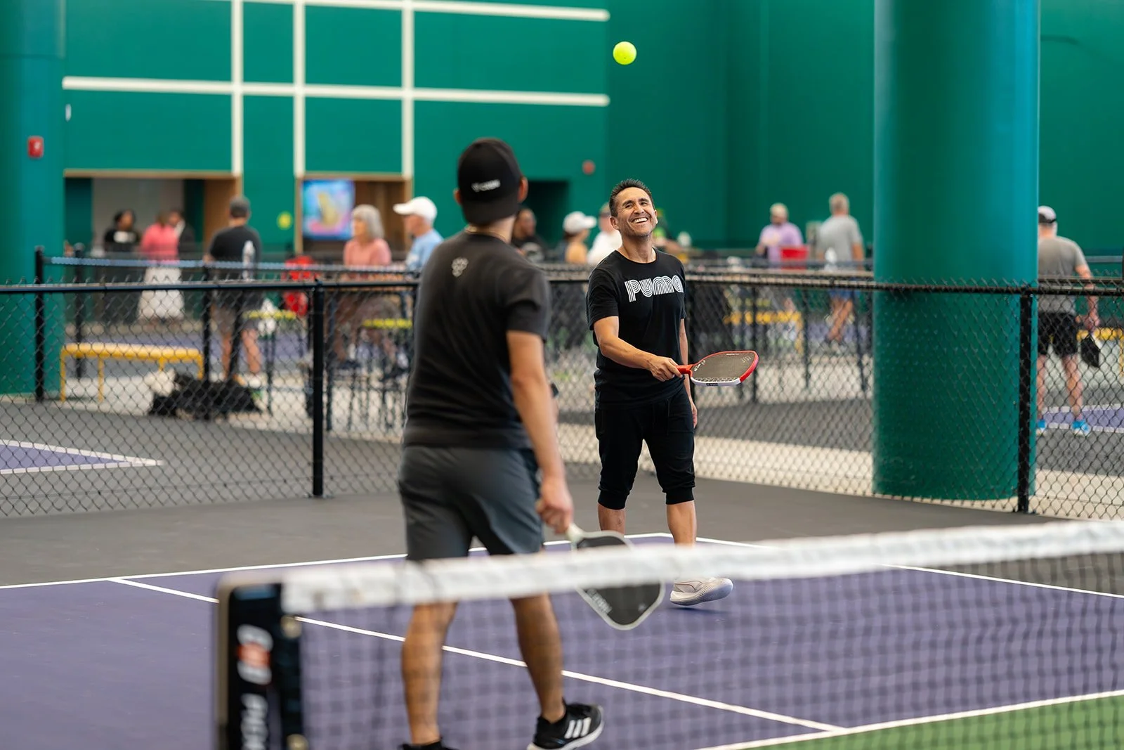 Two men playing pickleball on indoor court with a net, with multiple people in the background.