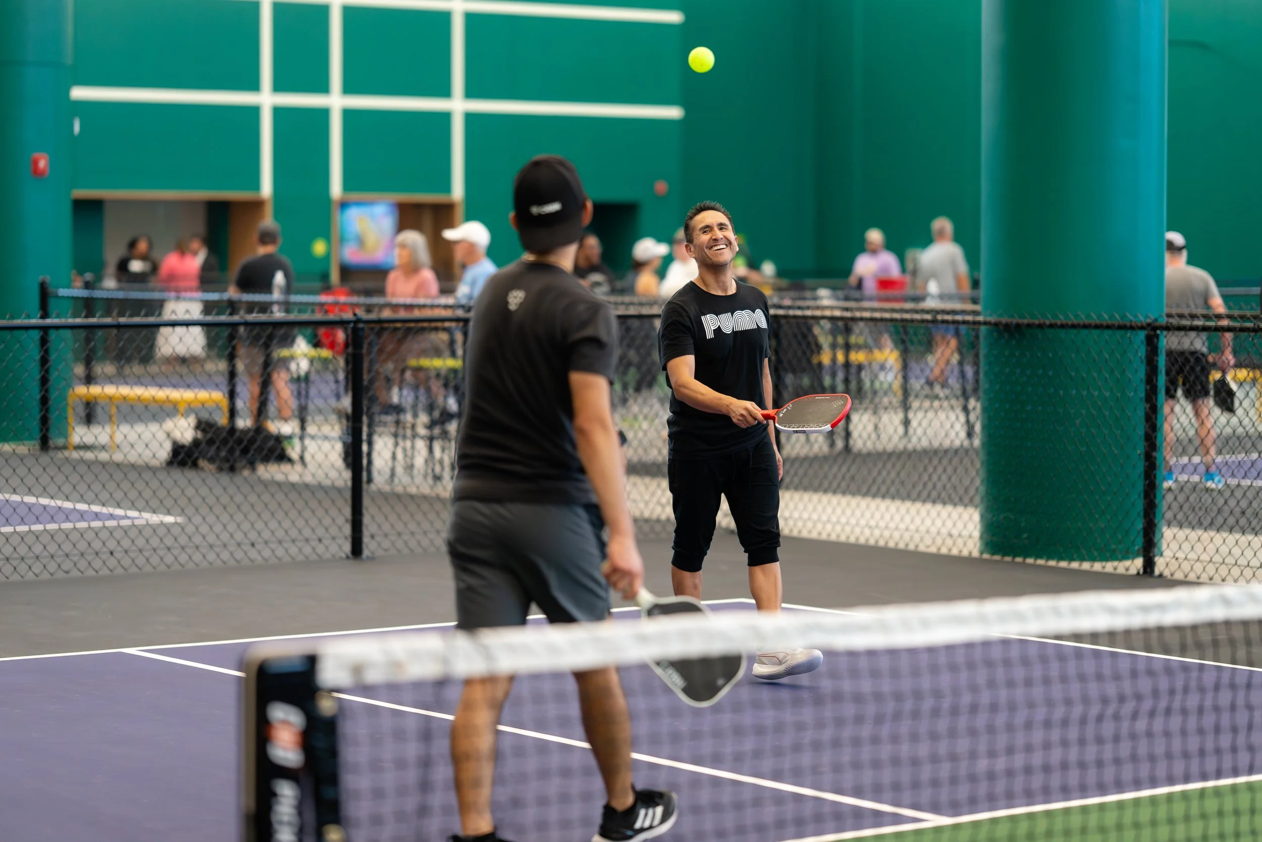 Two men playing pickleball on an indoor court, one smiling and holding a paddle, the other with his paddle at his side. Several people are in the background near a fenced area.