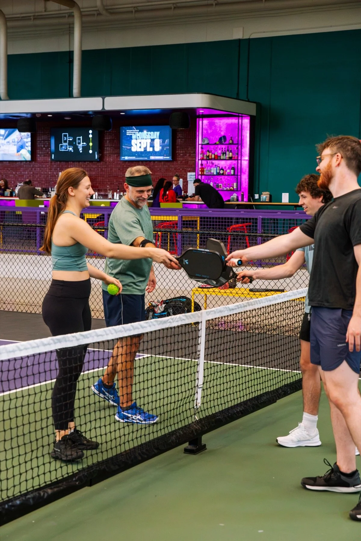 Indoor tennis courts with purple surfaces, surrounded by black fencing and green padding on the support columns, under a high wooden ceiling with large windows.