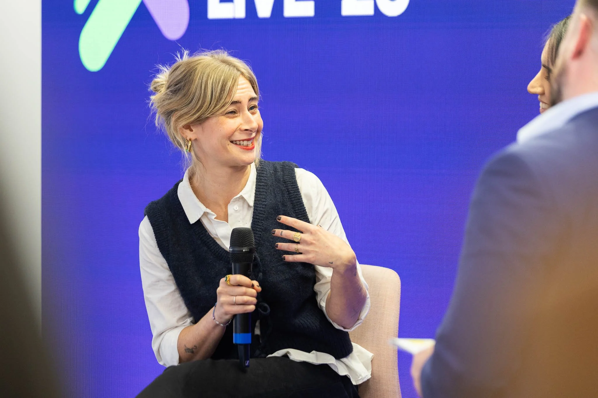 A woman with blonde hair styled in a loose bun, wearing a white shirt and dark vest, smiling and holding a microphone, engaged in conversation with a person wearing a gray blazer. The background features a large blue screen with partially visible text.