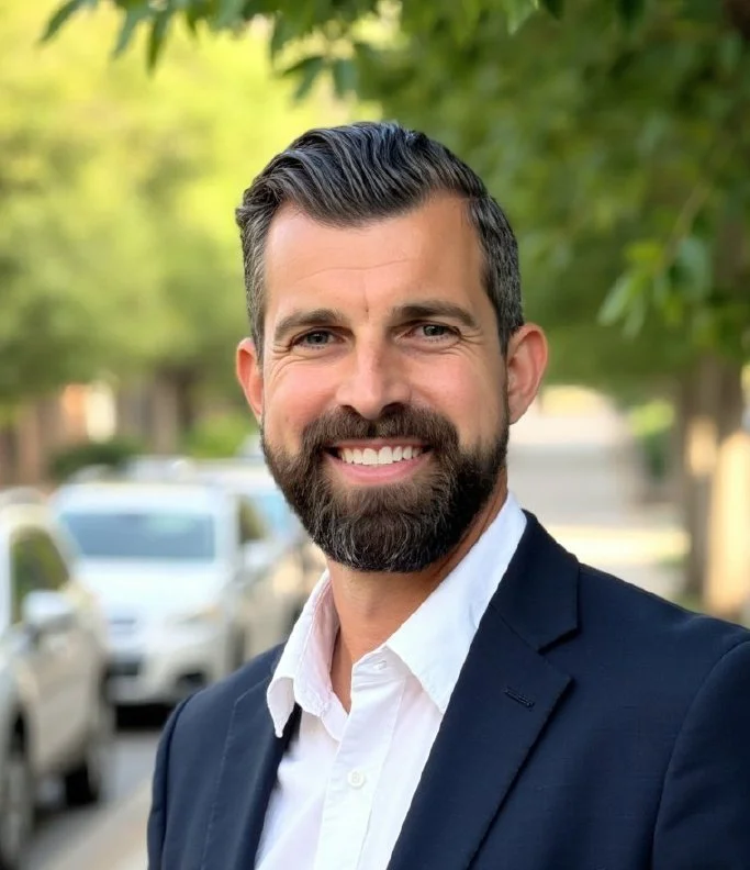 Portrait of a smiling man in a navy blazer and white shirt standing outdoors in a residential neighborhood with trees and parked cars in the background.