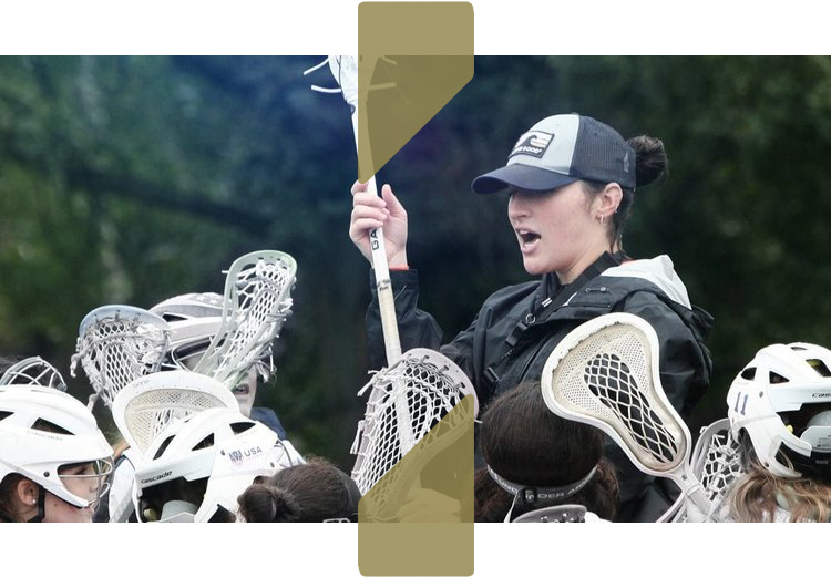 A female lacrosse coach in a cap gives a motivational talk to a group of players in helmets holding lacrosse sticks, outdoors.