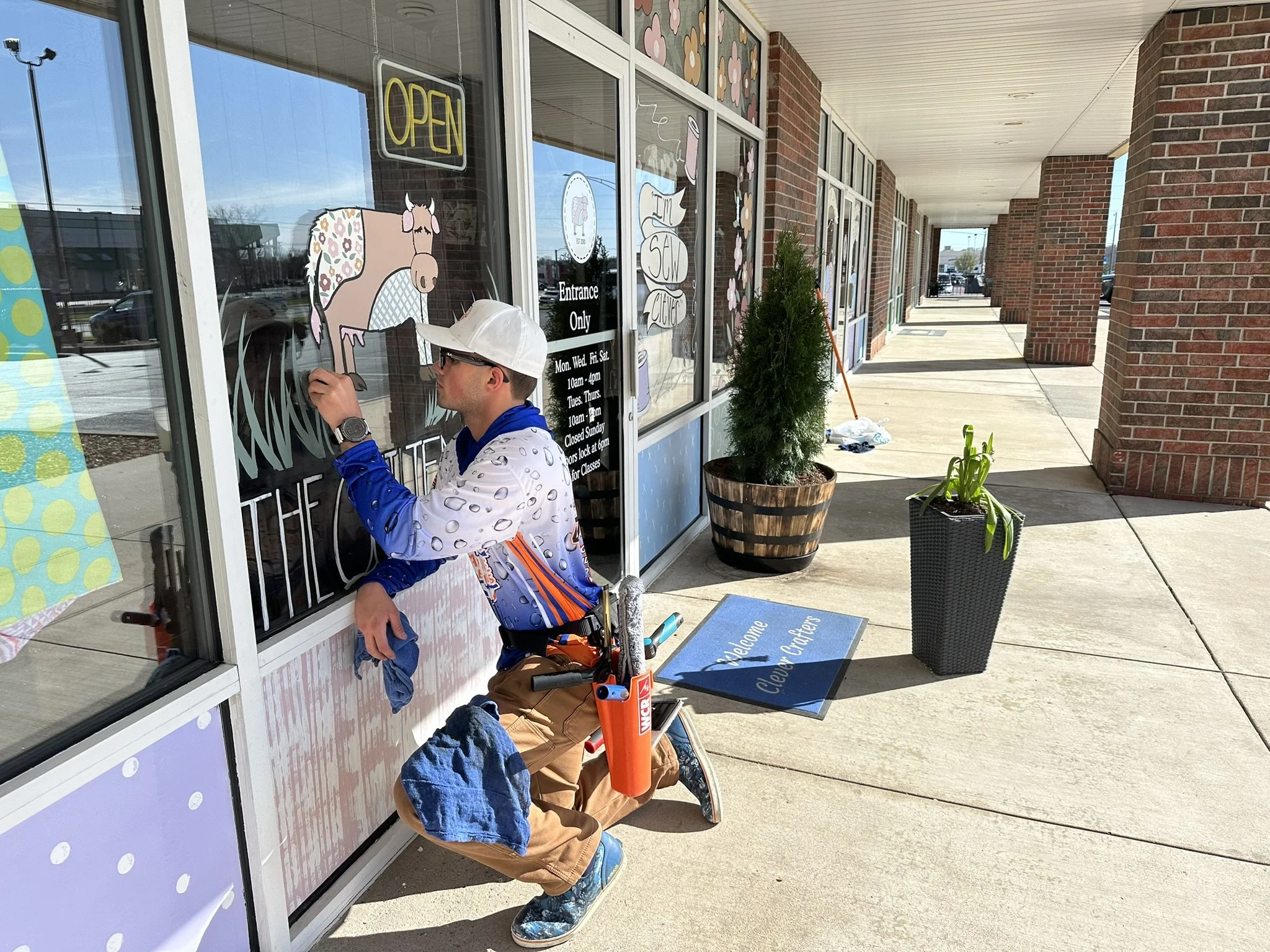 A person in a white cap, glasses, and a patterned jacket is painting a design on a storefront window. The storefront has an illuminated 'OPEN' sign and various decorations including a cartoon horse and floral patterns. There are potted plants outside on the sidewalk and a blue welcome mat that says 'Welcome Clever Cactus'.