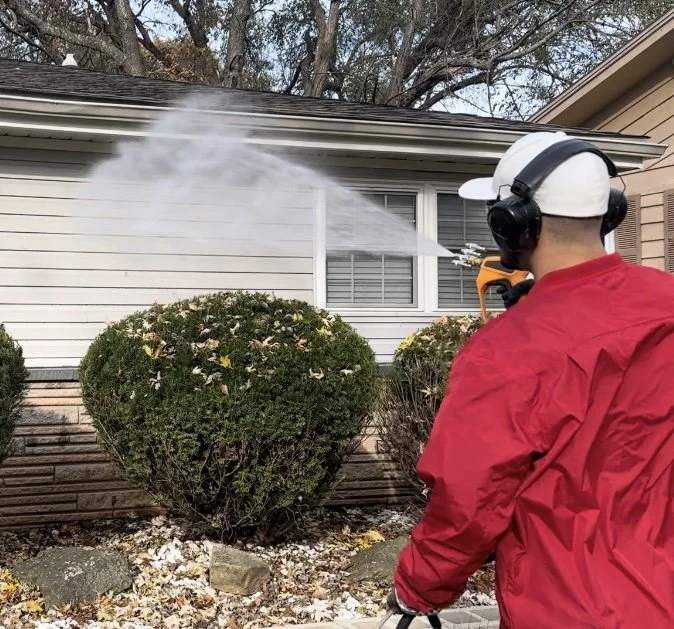 A person wearing a white hat, black headphones, and a red jacket using a power washer to clean bushes in front of a house.