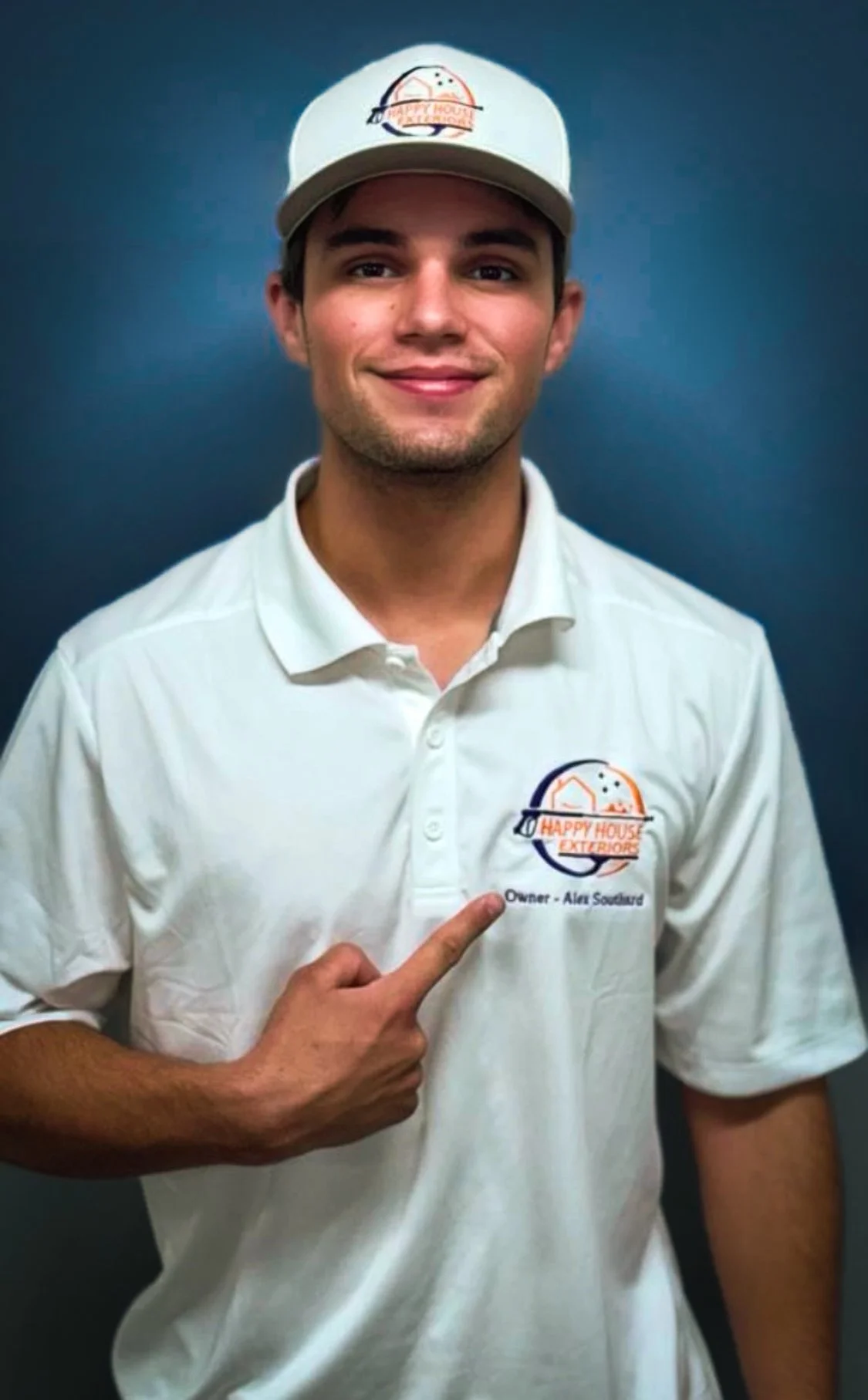 Young man wearing a white polo shirt and cap pointing to the logo on his shirt that reads 'Happy House Exterior' with the owner’s name ‘Ali Saad’ underneath, against a dark background.