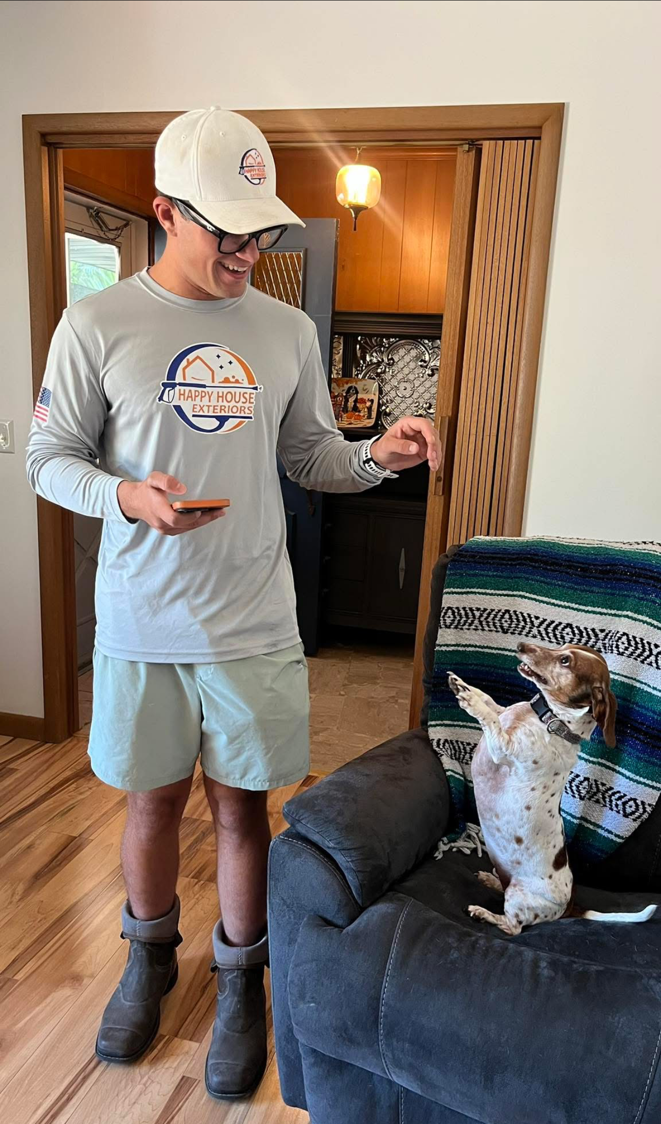 A young man wearing glasses, a baseball cap, a long-sleeve shirt, and shorts standing next to a dog on a couch. The man is smiling and holding a smartphone, while the dog is sitting on the couch with front paws raised, looking at the man.