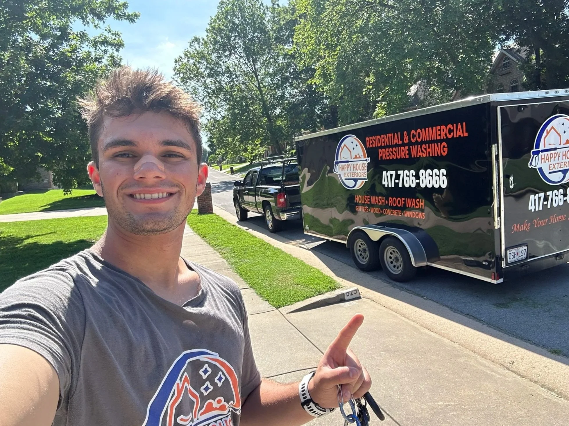 A young man smiling and taking a selfie outdoors on a sunny day, pointing at a black service trailer with red and white text that reads 'Happy House Exteriors' and advertises residential and commercial pressure washing services.