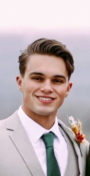 A young man smiling outdoors, wearing a light gray suit, white shirt, and green tie, with a small floral boutonniere on his lapel.