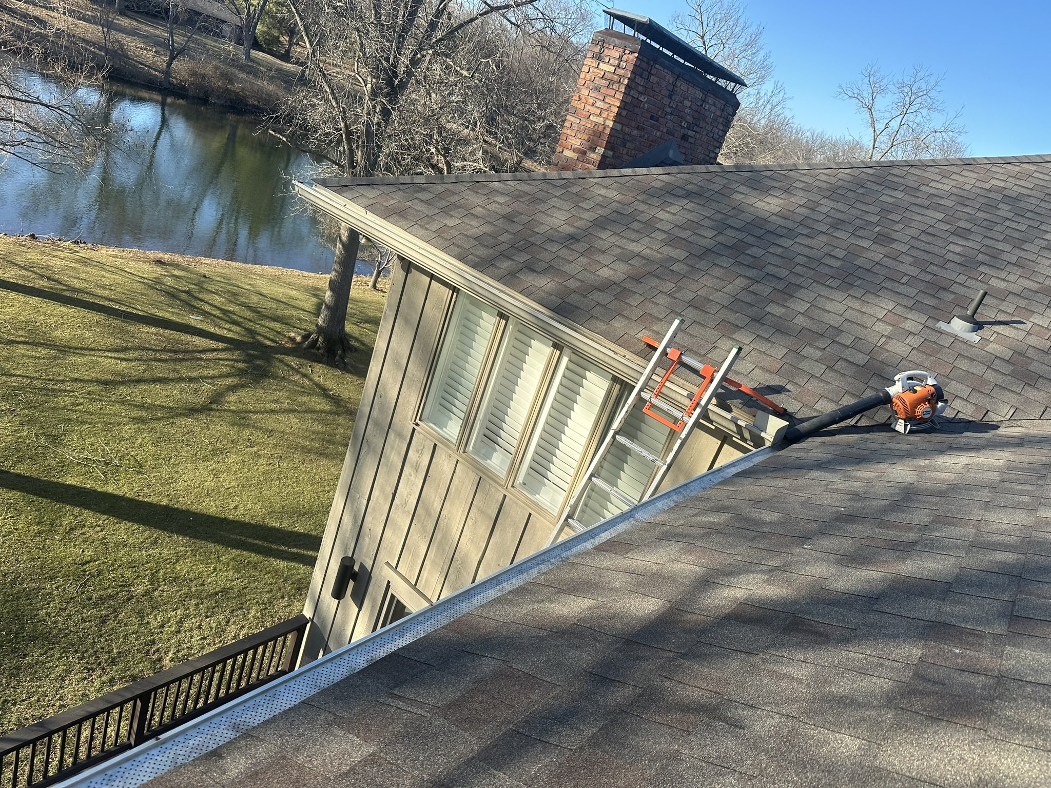 A house roof with a ladder and leaf blower, overlooking a yard with a tree and a pond in the background.