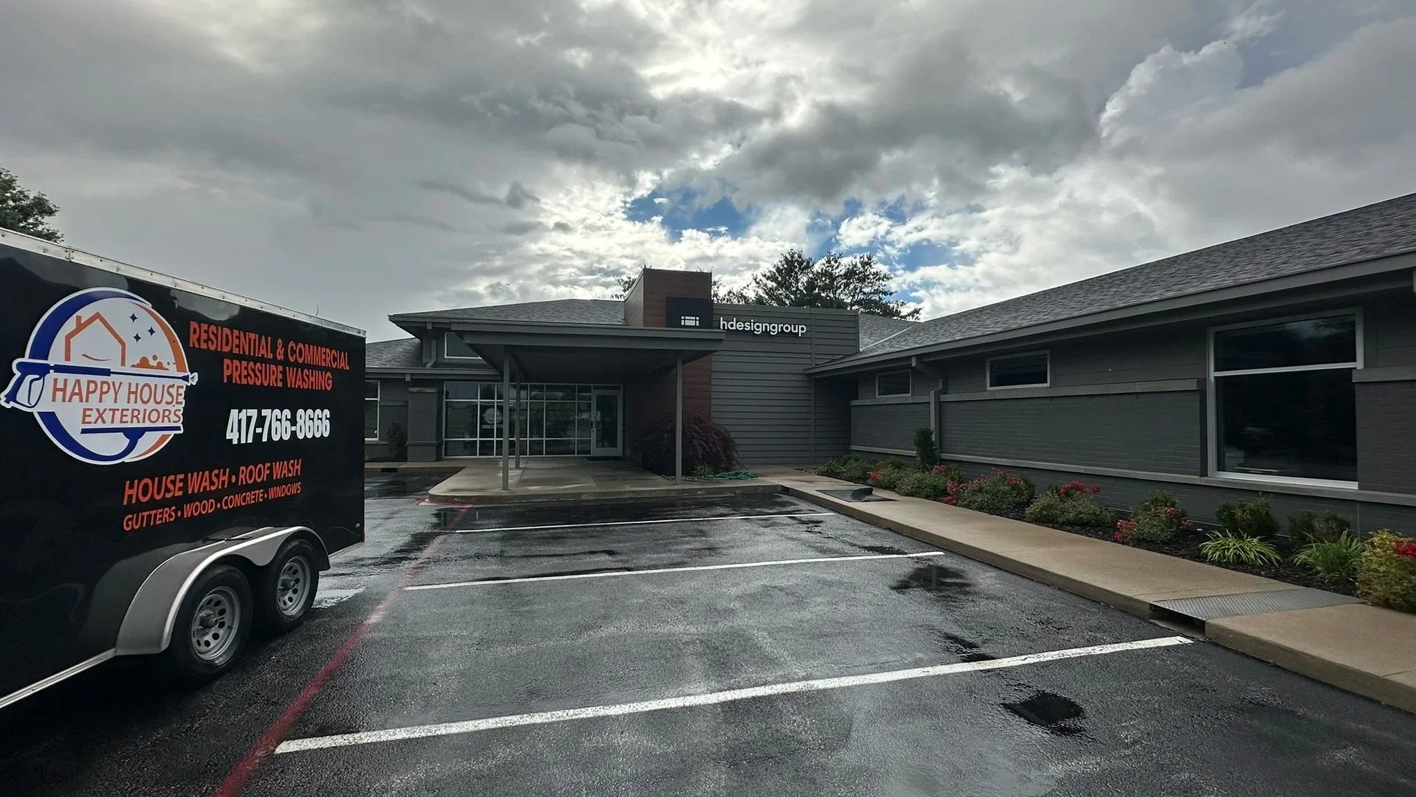 A commercial building with a sign for HDESIGN GROUP on the front, and a trailer for Happy House Exteriors offering residential and commercial pressure washing services, including house wash, roof wash, gutters, wood, concrete, and windows, with a parking lot in front showing wet pavement and parking spaces outlined in white.