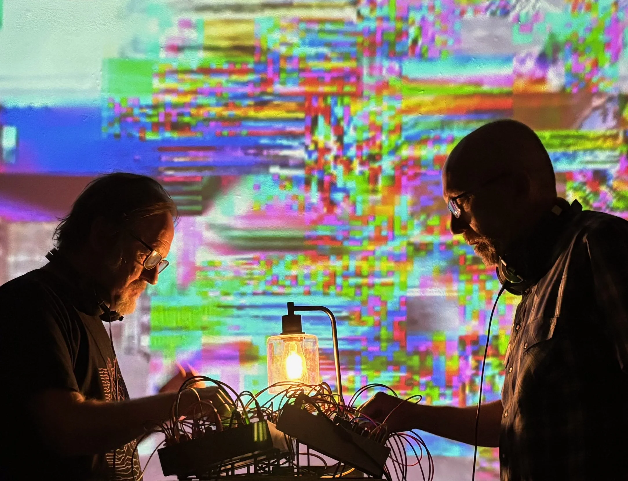 Two people with long hair and glasses are seen working at a table with electronic equipment, with a colorful, pixelated digital background projected behind them.