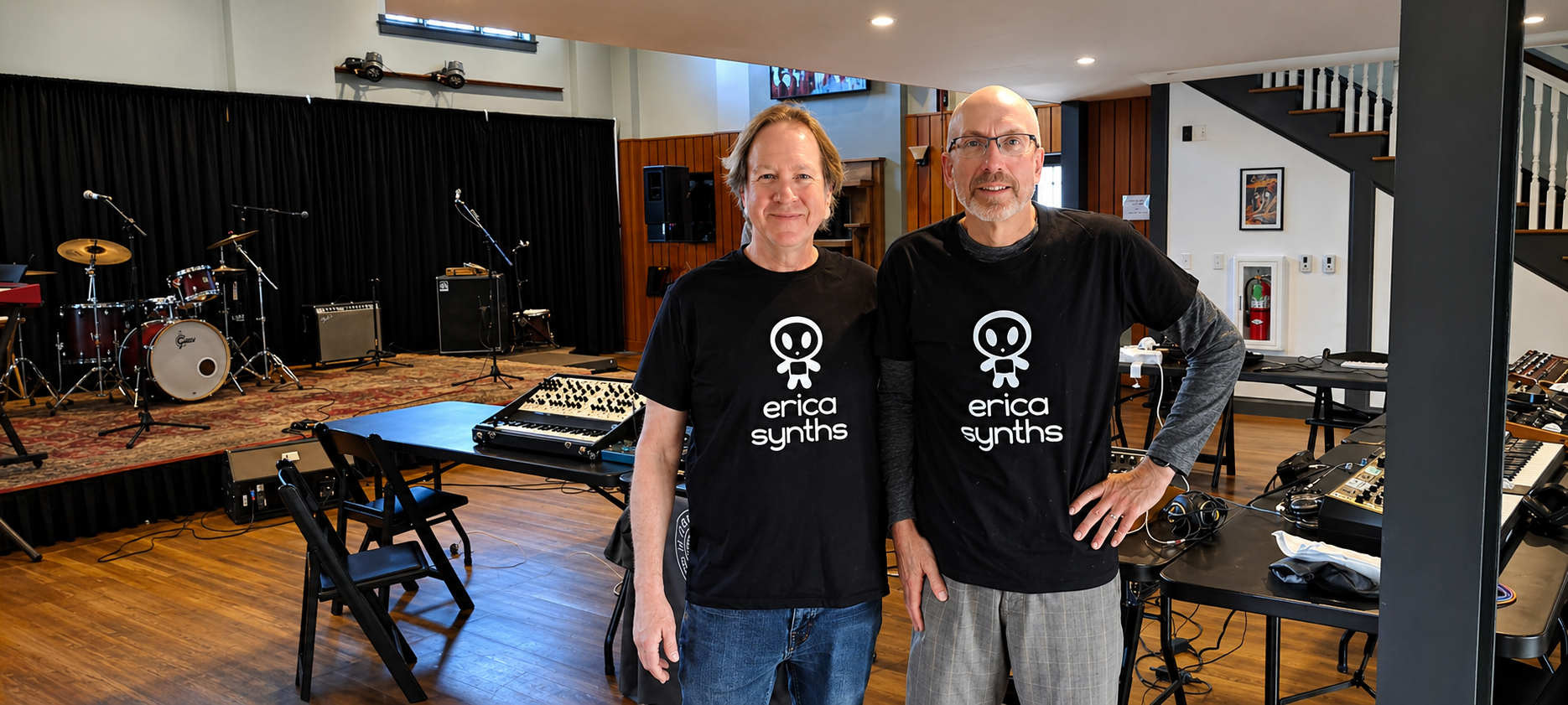 Two men wearing black t-shirts with a skull logo and 'erica synths' text, standing in a music rehearsal space with drums, keyboards, and audio equipment.
