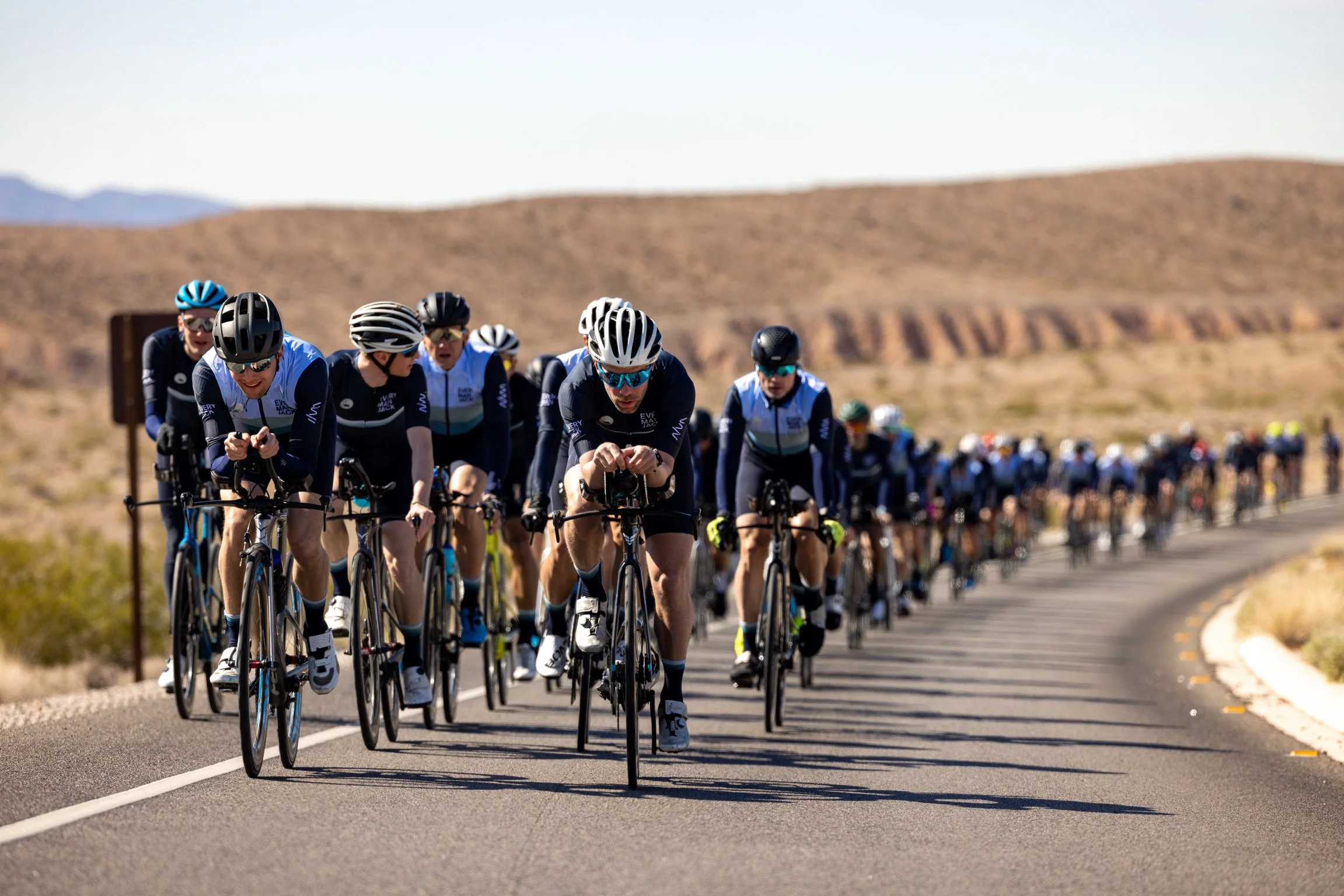 A large group of cyclists participating in a cycling event on a road in a desert landscape with mountains in the background.