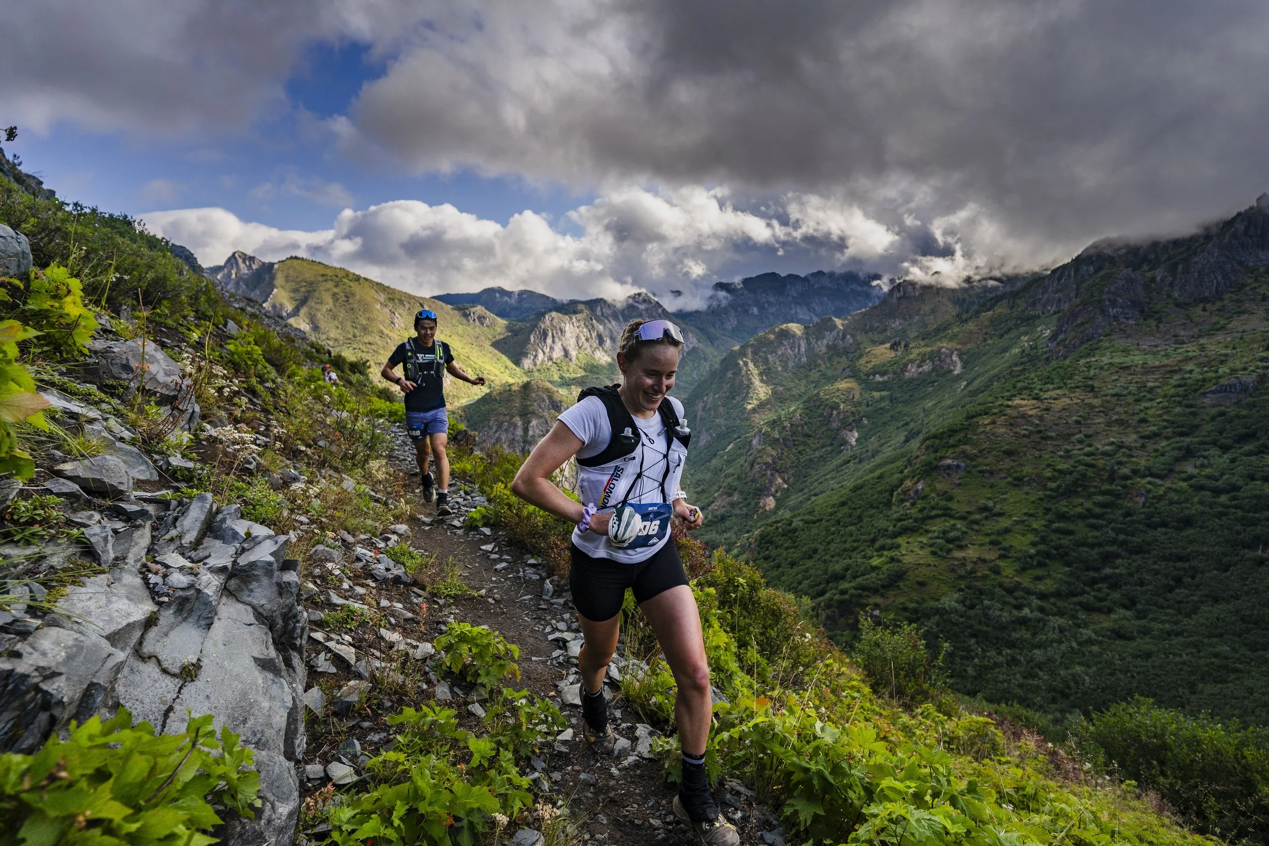 Two runners, a man and a woman, trail running on a narrow mountain path surrounded by green vegetation with mountains and a cloudy sky in the background.