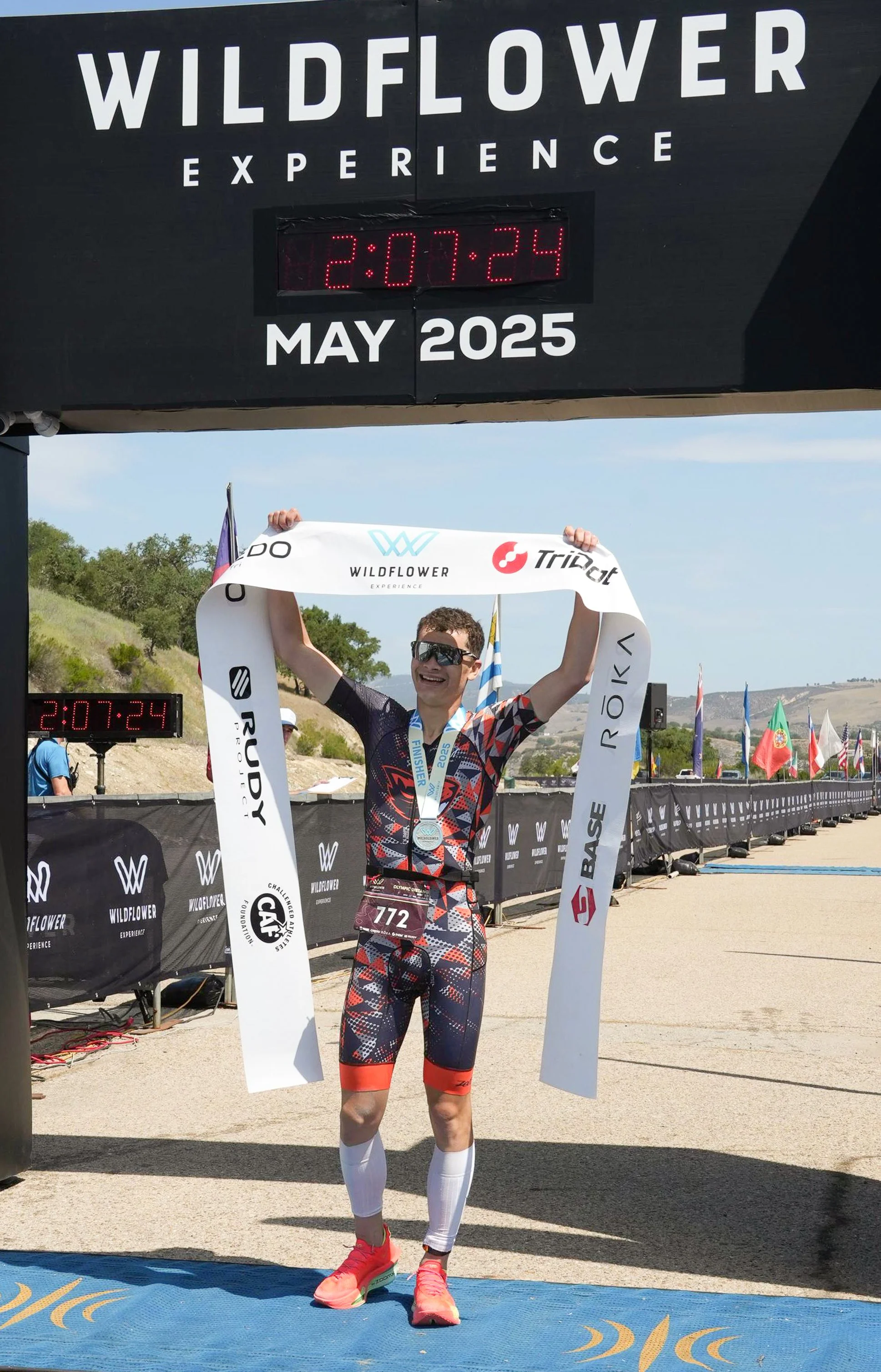 A male marathon runner is celebrating crossing the finish line at the Wildflower Experience race in May 2025, holding up the finish line tape and wearing a medal. The digital clock shows the time as 2:07:24.