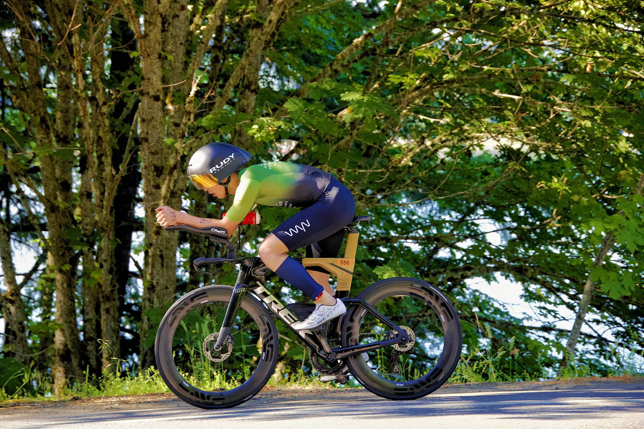 Cyclist in green and black outfit riding a black Trek time trial bike with disc wheels on a roads surrounded by green trees.