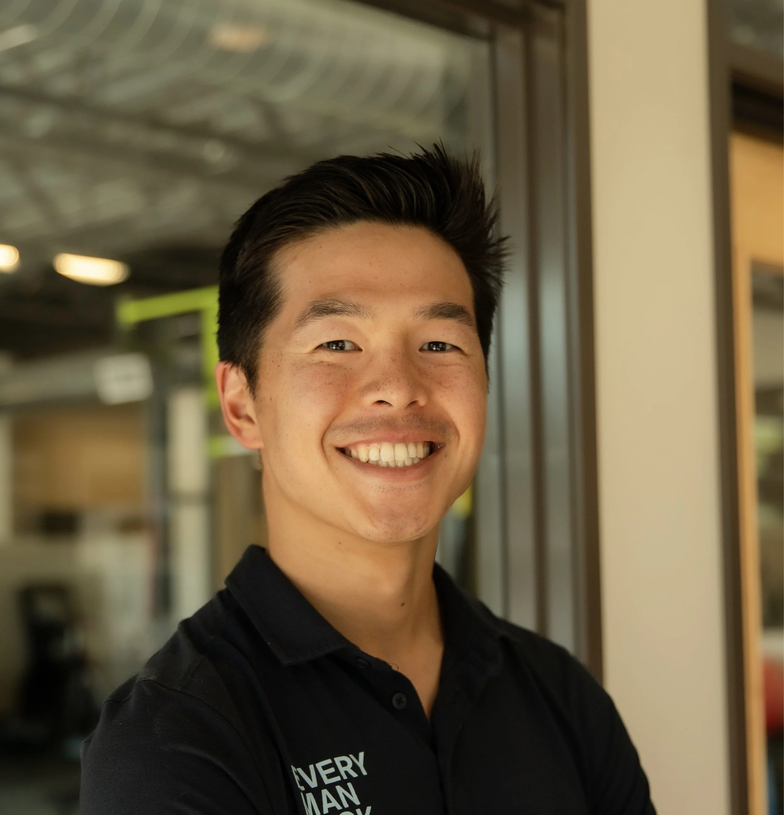 Smiling young man with short black hair wearing a black polo shirt indoors.