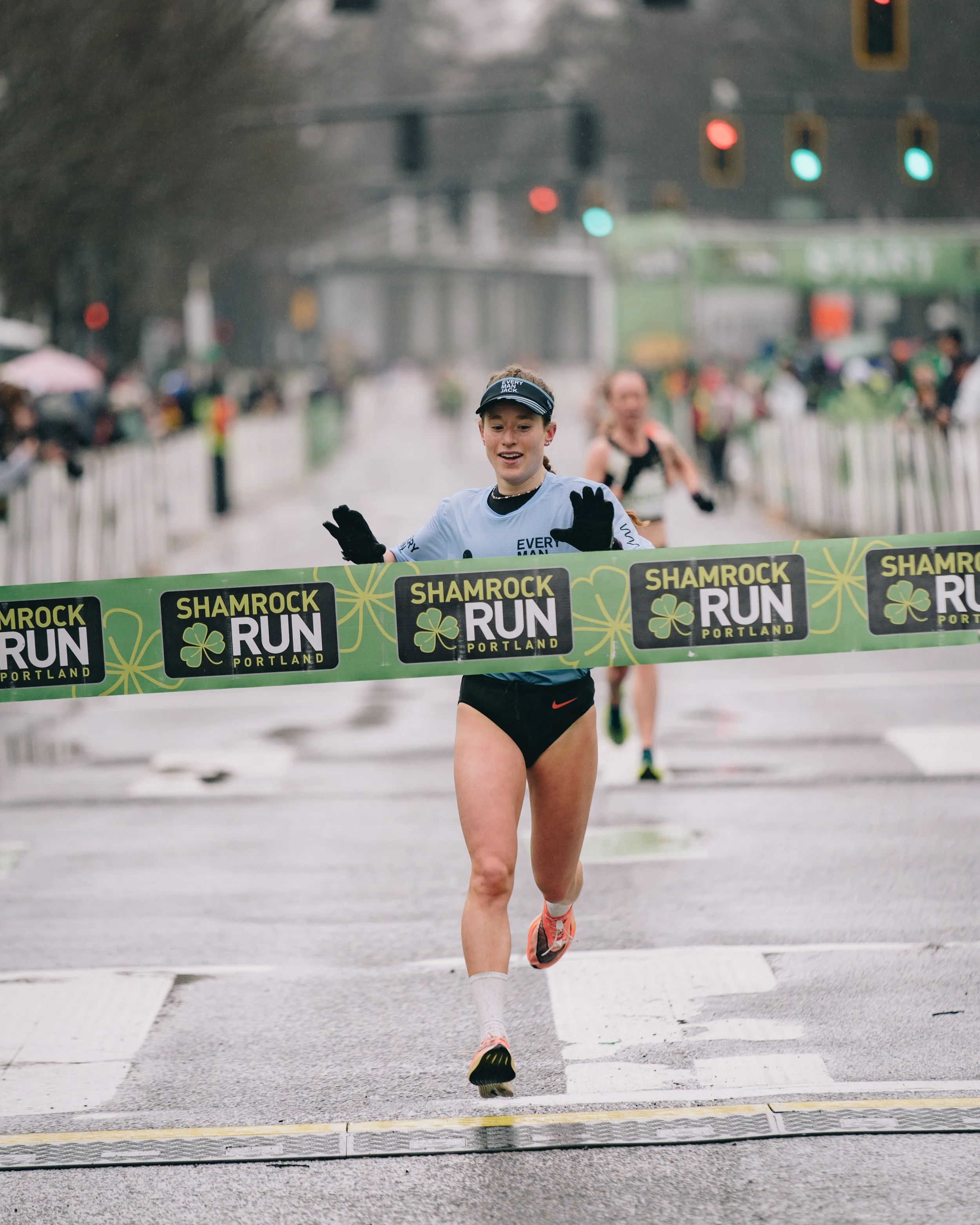 A female runner crossing the finish line at the Shamrock Run Portland, with a joyful expression. She is wearing a light blue shirt, black shorts, and a black visor. The race is in rainy weather, with water on the ground and spectators behind barriers holding umbrellas.