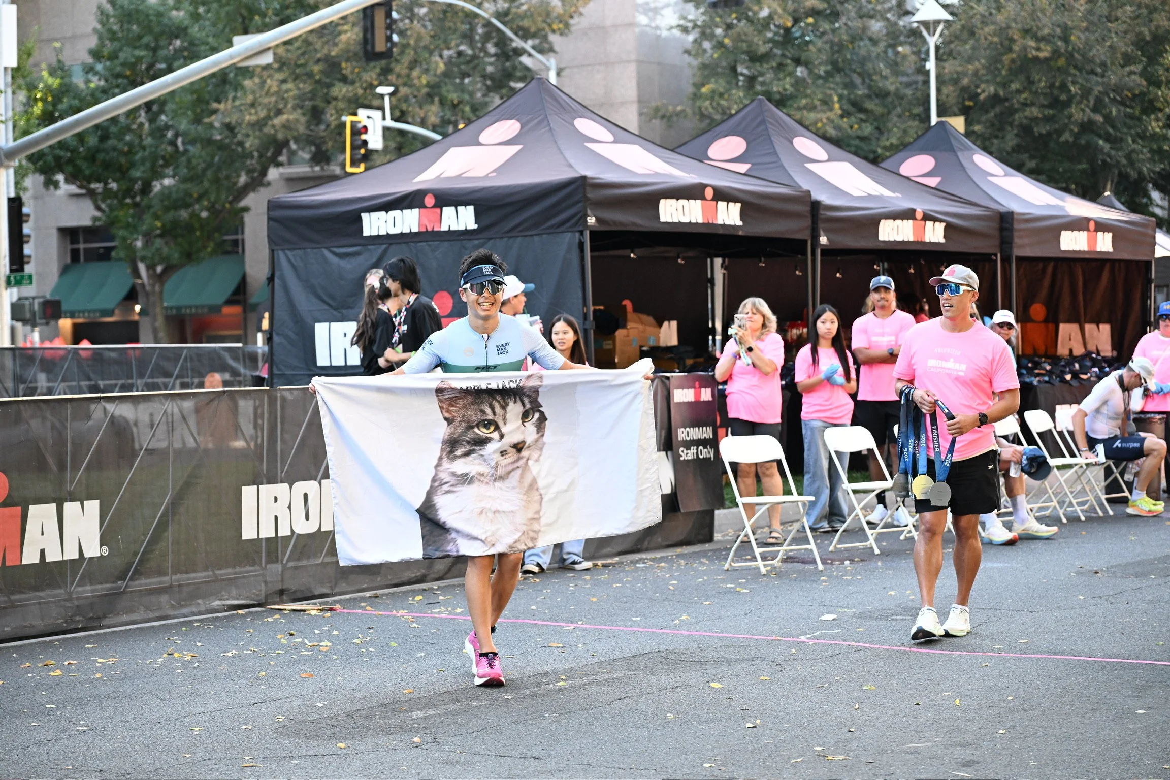 A man running in a marathon holding a large flag with a cat image, surrounded by event volunteers and spectators in pink shirts at an IRONMAN race.