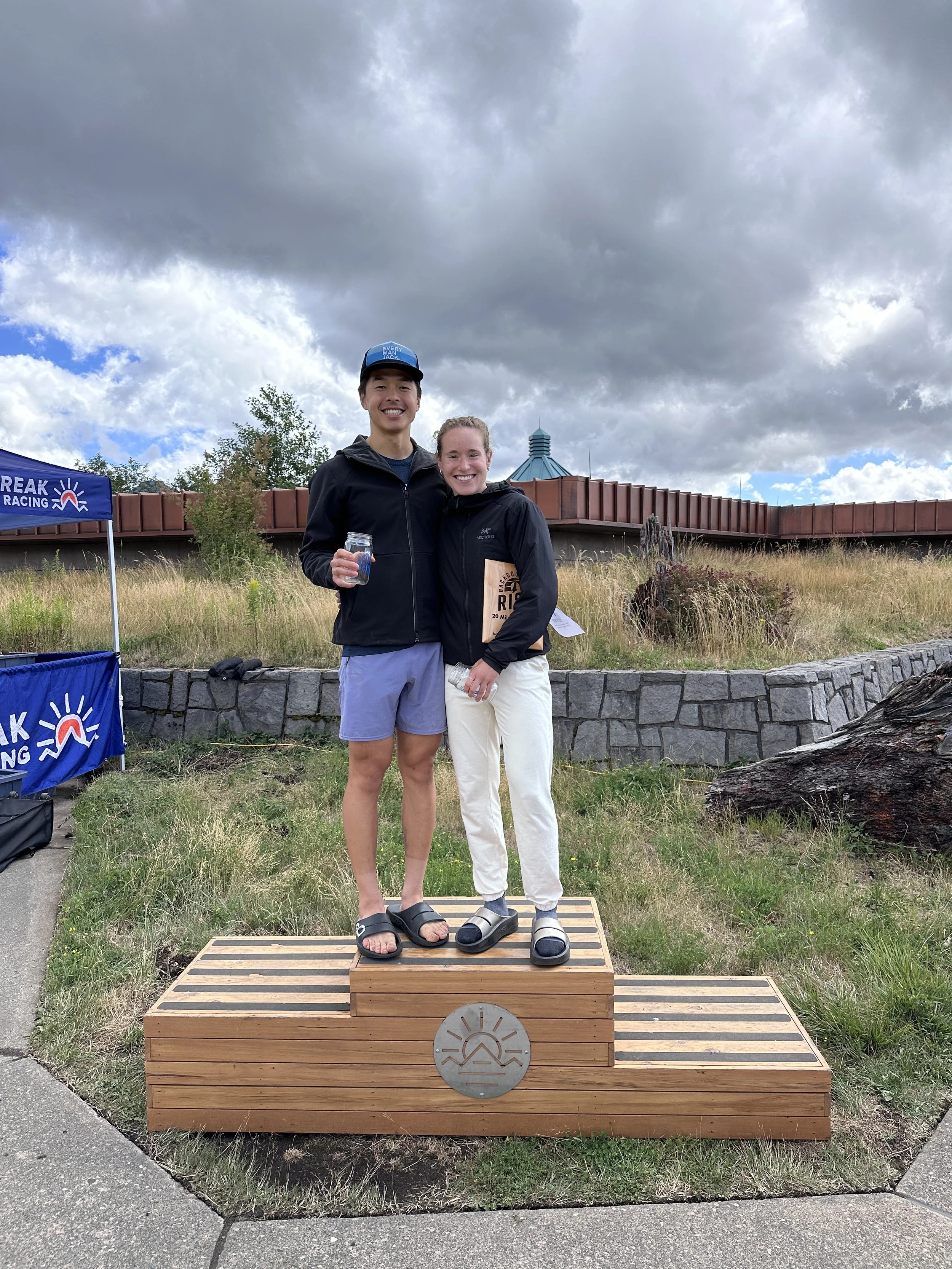 Two young adults, a man and a woman, standing on a wooden podium outdoors, smiling at the camera, with the man holding a glass jar. The man is wearing a black jacket, light blue shorts, and a blue cap, while the woman is dressed in a black jacket and white pants. They are standing in front of a grassy area with a stone wall, ornamental grass, and a building with a dome and a cloudy sky in the background.