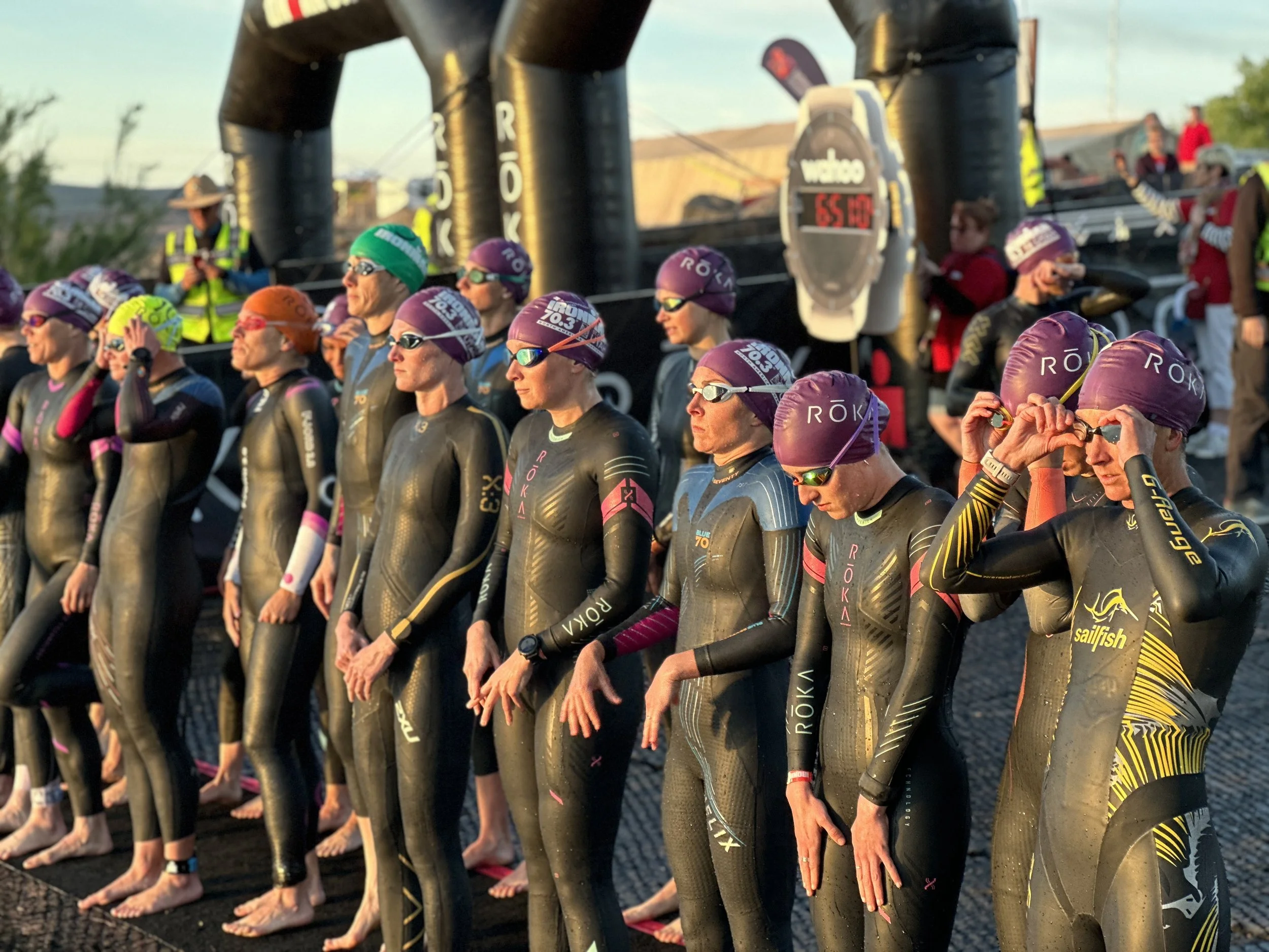 A group of athletes in wetsuits and swim caps standing at the starting line of a race, with some adjusting goggles and focusing ahead. The setting appears to be outdoors during sunrise or sunset. Ironman