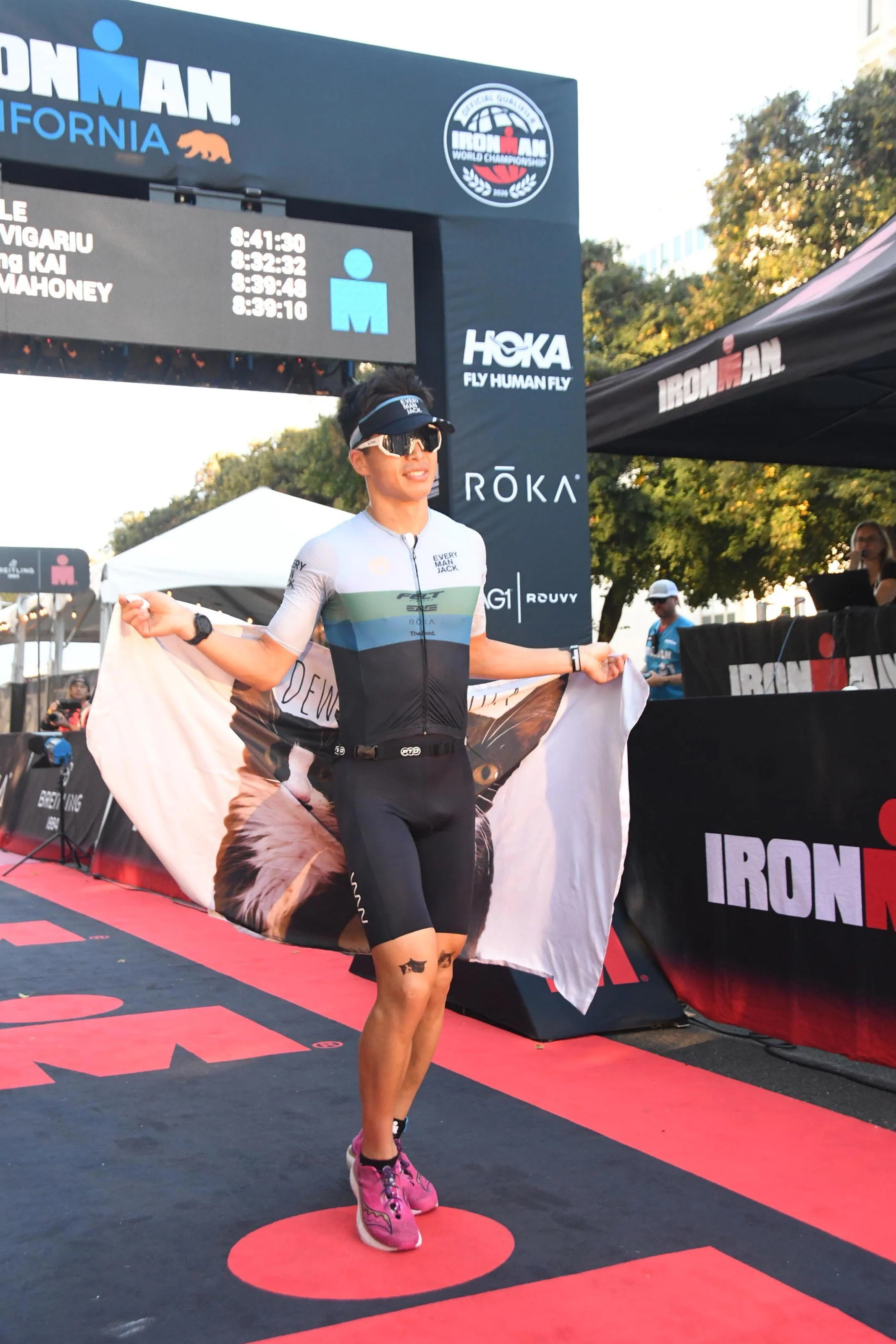A male triathlete celebrating crosses the finish line of an Ironman race, holding a flag in a stadium with a large digital timer and banners in the background.